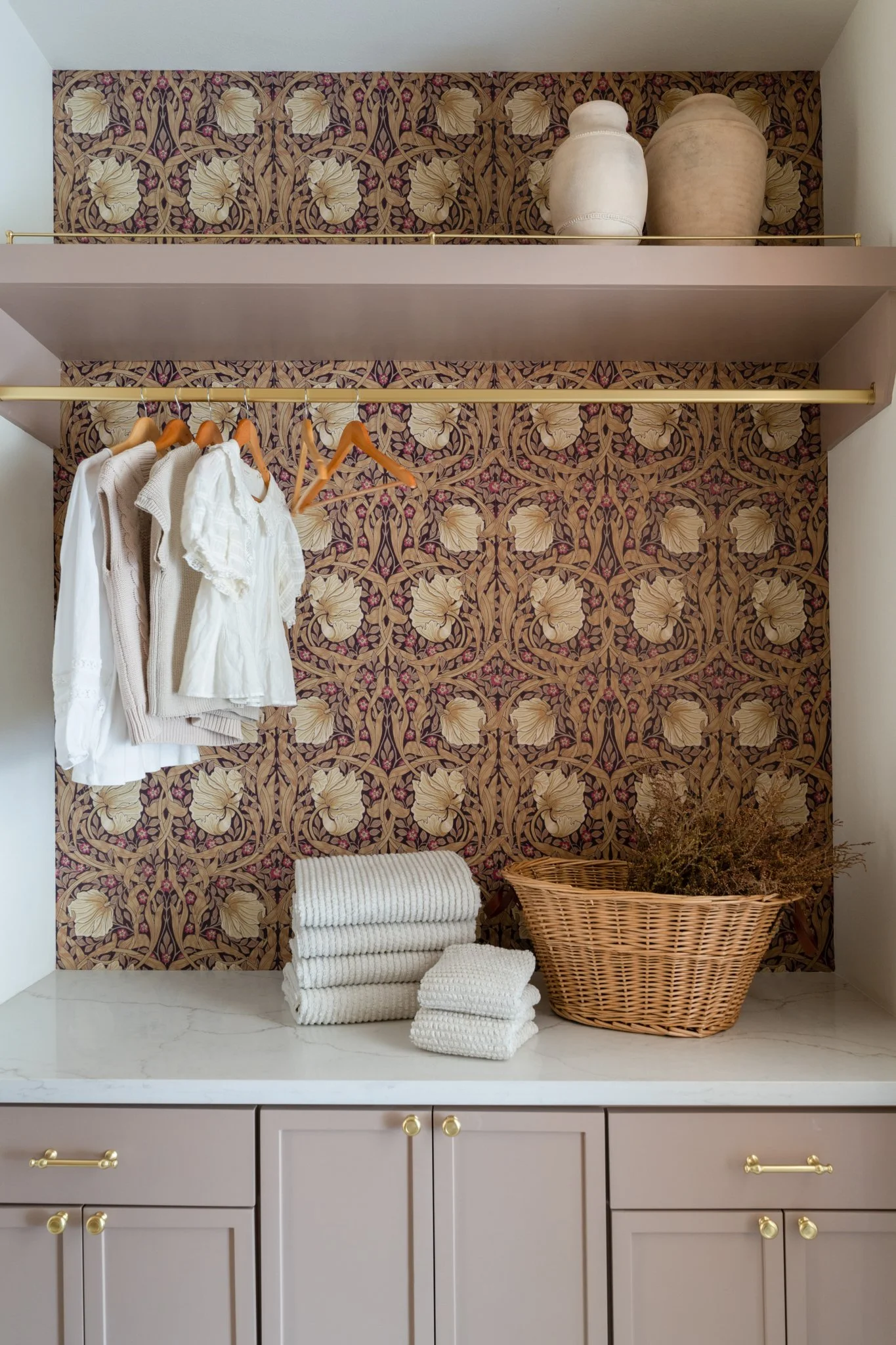 Closet with decorative wallpaper, hanging clothes, folded towels, basket with dried plants, and decorative vases on upper shelf.