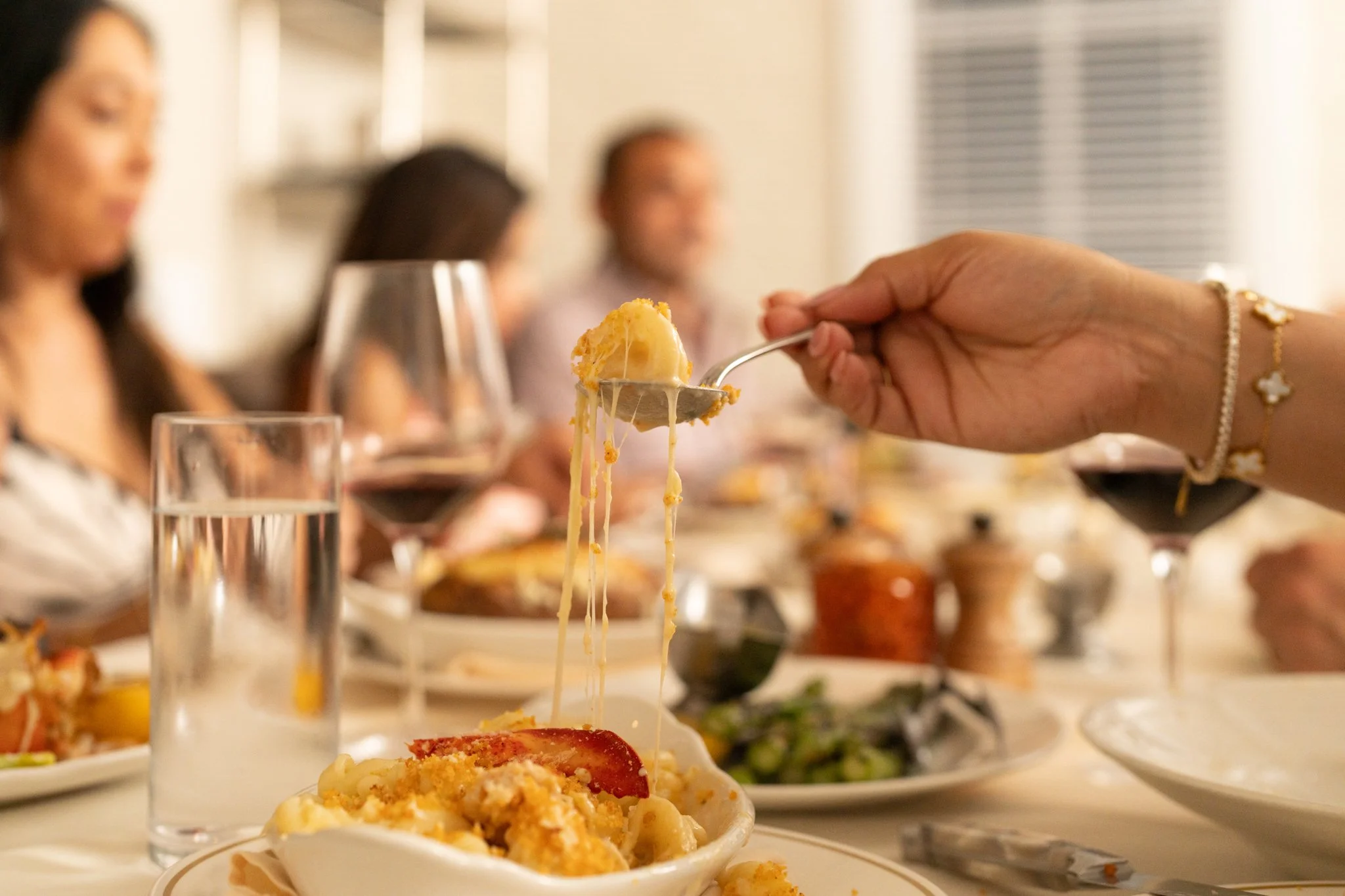 Restaurant Photography of Dean's Steakhouse at Kimpton Santo in San Antonio  A person holding a spoonful of cheesy casserole over a plate of food at a dinner party with people and wine glasses in the background.