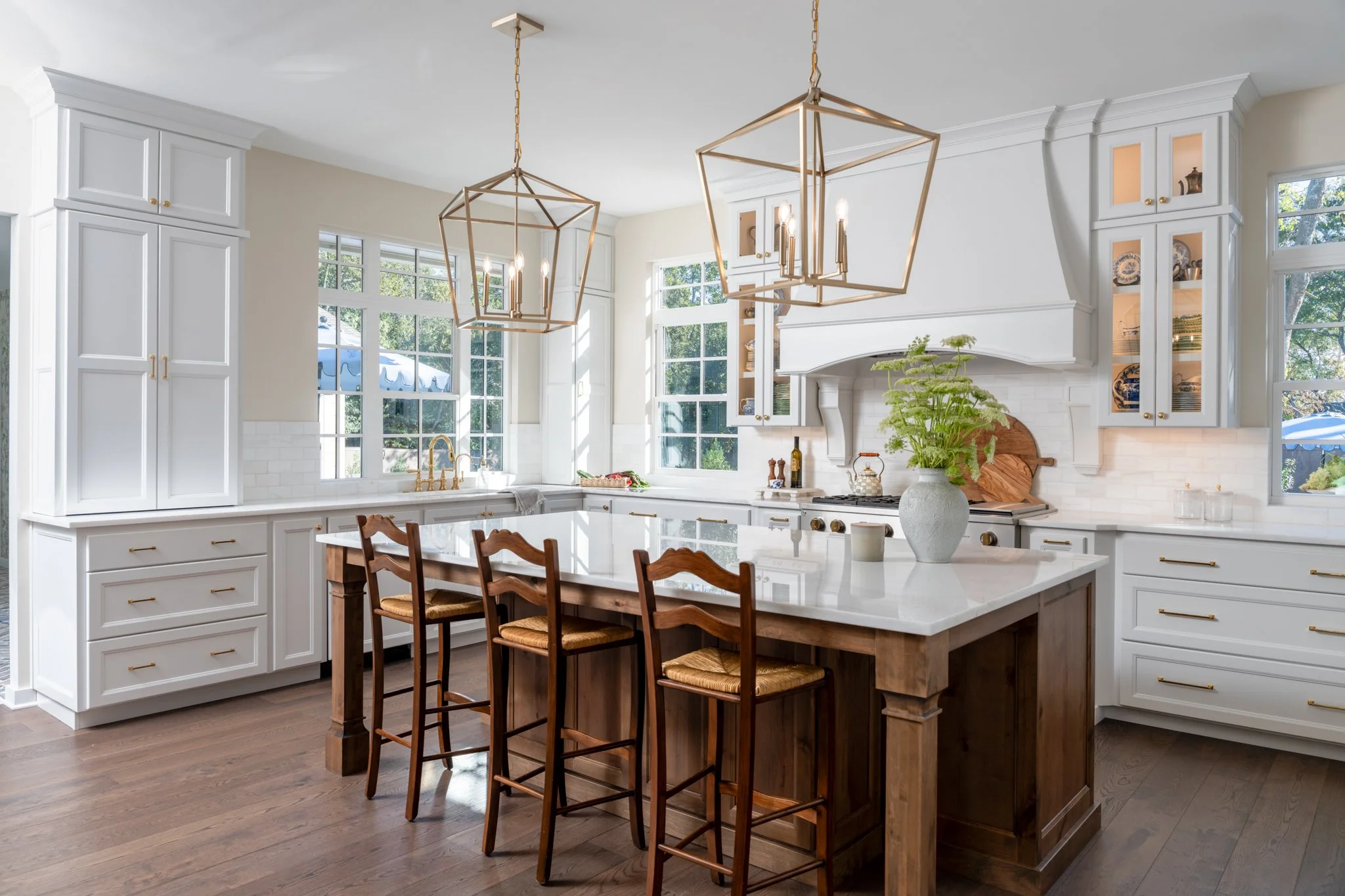 Bright kitchen with white cabinets, large windows, and a central island with a wooden base and white countertop. Four wooden bar stools with woven seats are at the island. Two pendant lights hang from the ceiling, and there is a large vase with green