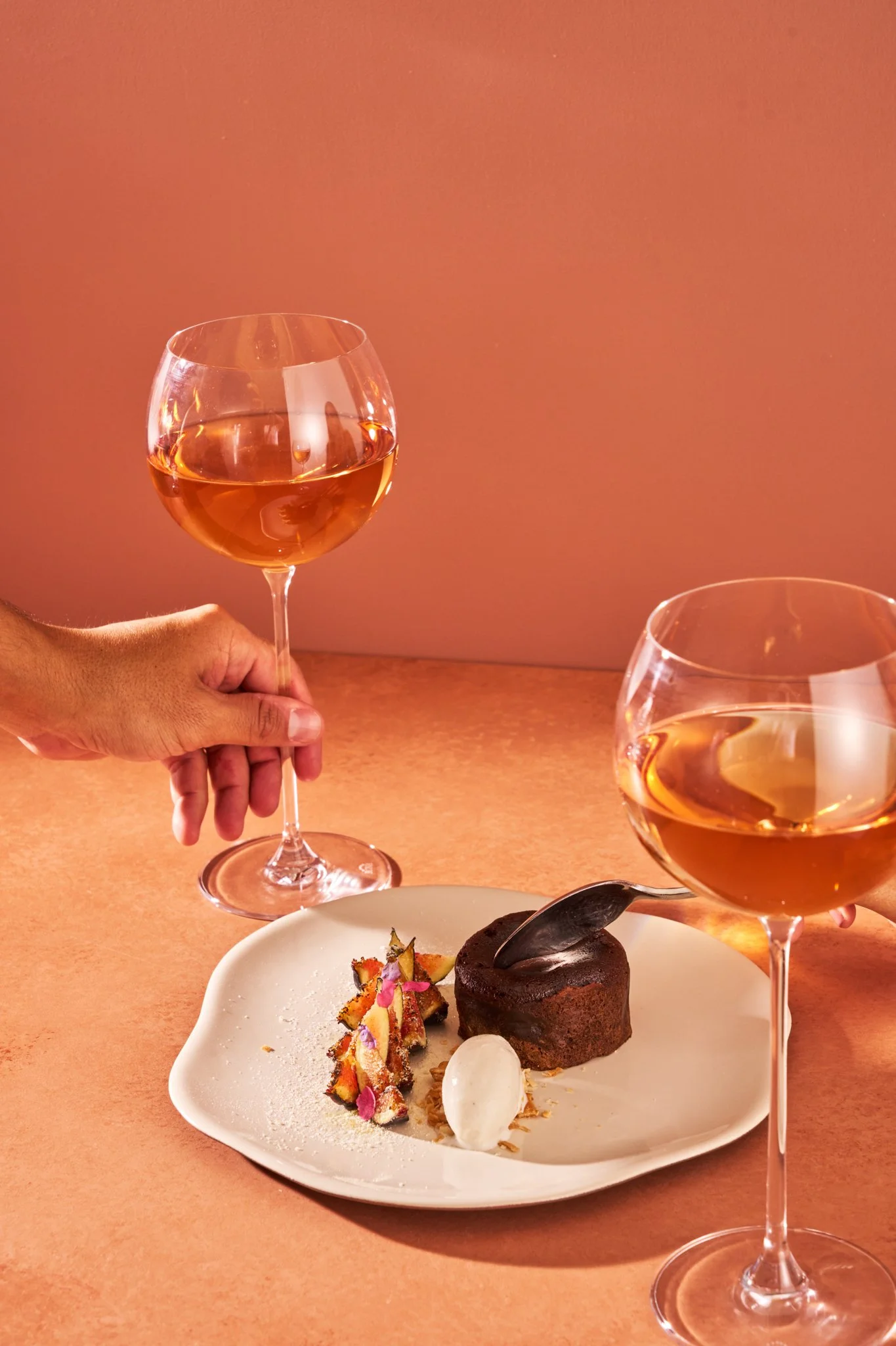 Food Photography featuringa dessert plate with a chocolate lava cake, a scoop of vanilla ice cream, and decorative fruit garnishes, served with two glasses of rosé wine on a pink background by Richard Sandoval Hospitality Group in Denver, CO