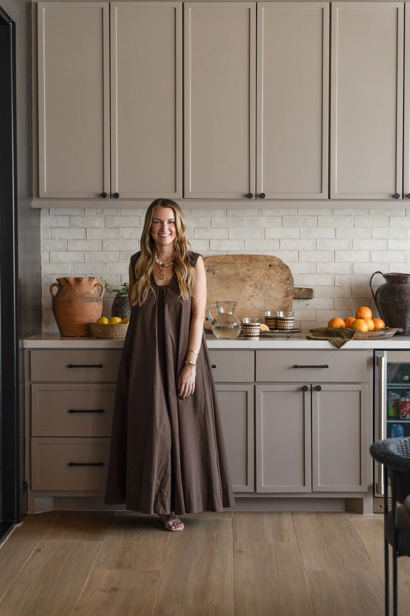 A woman in a long brown dress standing in a kitchen with beige cabinets, a brick backsplash, and various kitchen items on the counter.