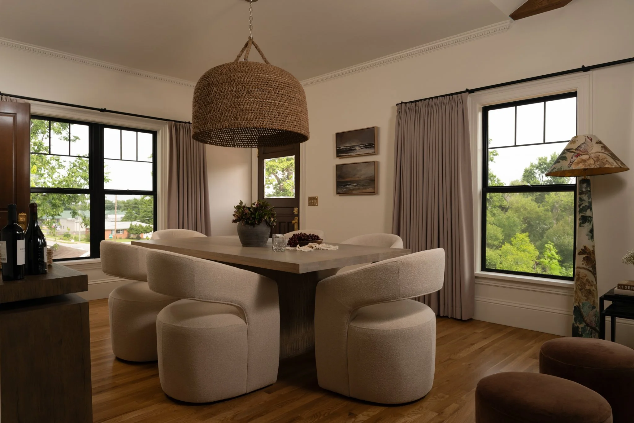 Dining room with large windows, beige upholstered chairs around a wooden table, a wicker pendant light, and a floor lamp with a patterned shade.