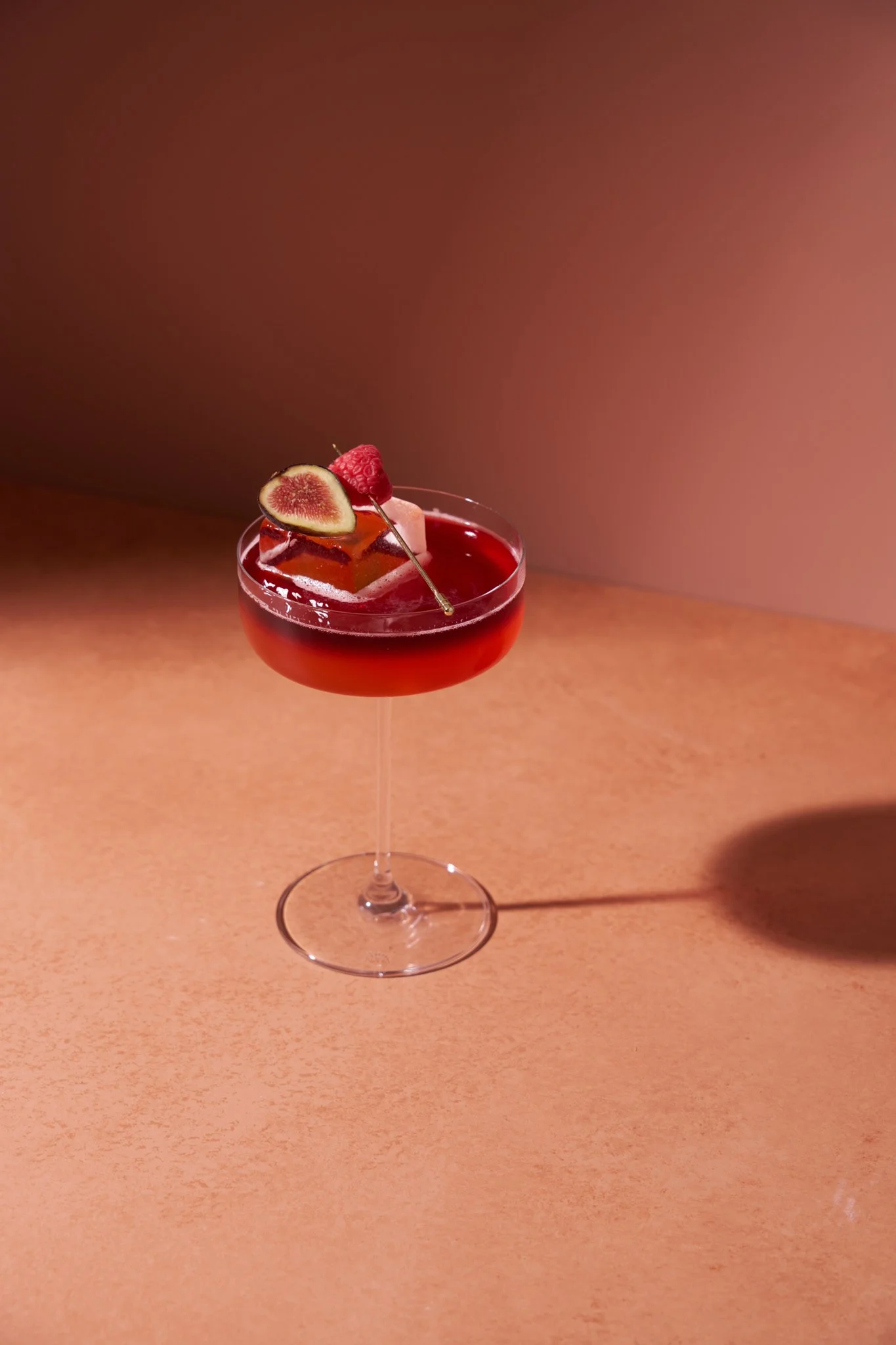 Beverage photography  for Richard Sandoval Hospitality featuring  A cocktail in a coupe glass with red liquid, garnished with a slice of fig, a raspberry, and a pink square-shaped ice cube, on a light brown surface with shadows.