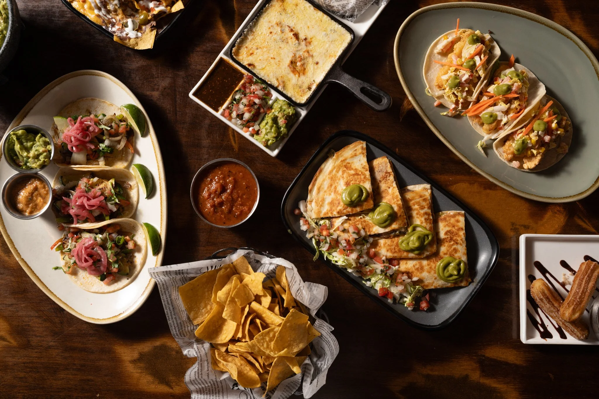 Food photography for Richard Sandoval restaurant Venga Venga in Aspen, CO of A variety of Mexican dishes on a wooden table, including tacos, quesadillas, nachos with salsa and guacamole, churros, and fajitas, accompanied by sauces and lime wedges.