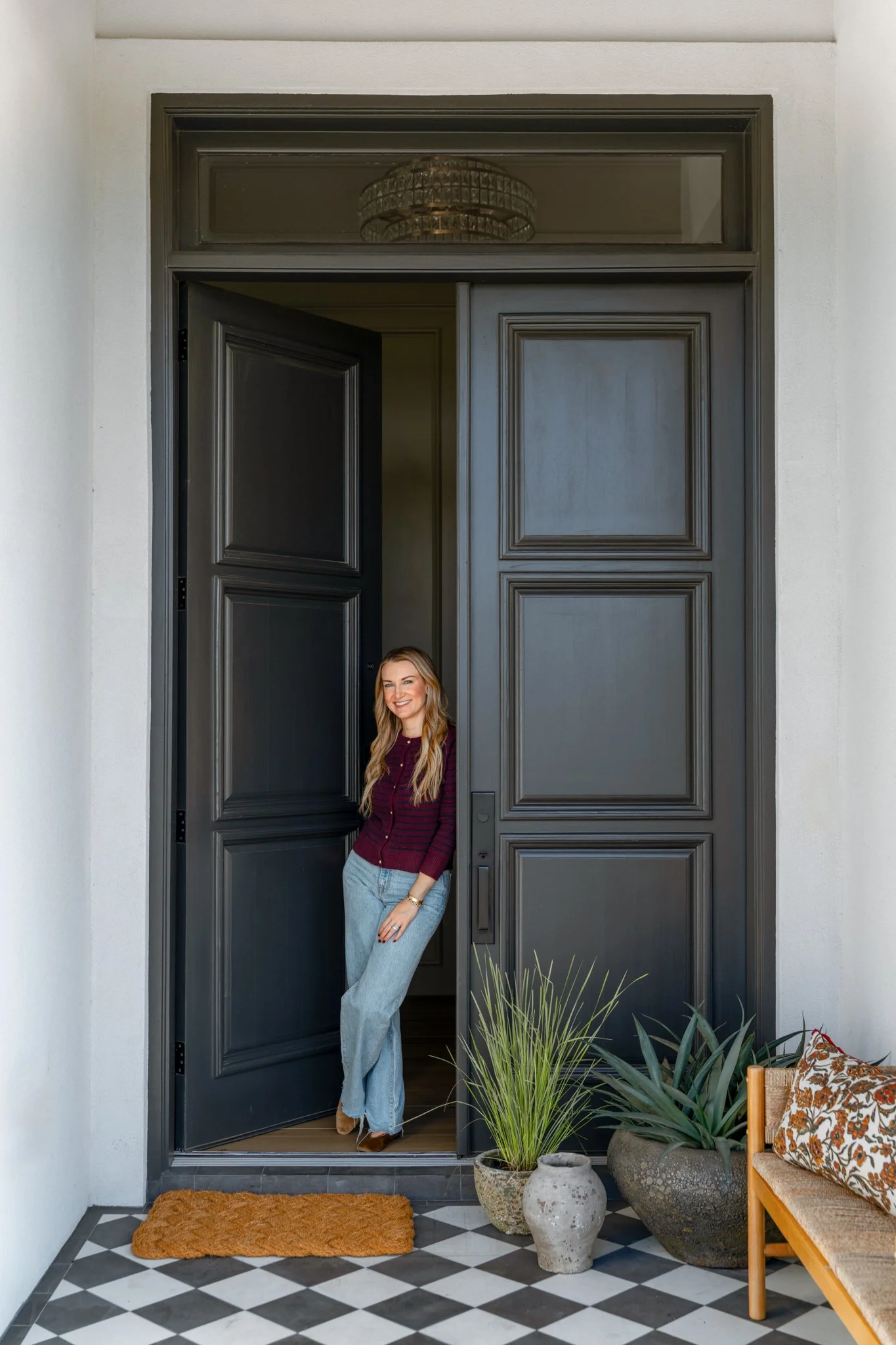 A woman standing in the doorway of a home, partially outside, with black double doors and a white wall, plants, and a bench with a floral cushion nearby.