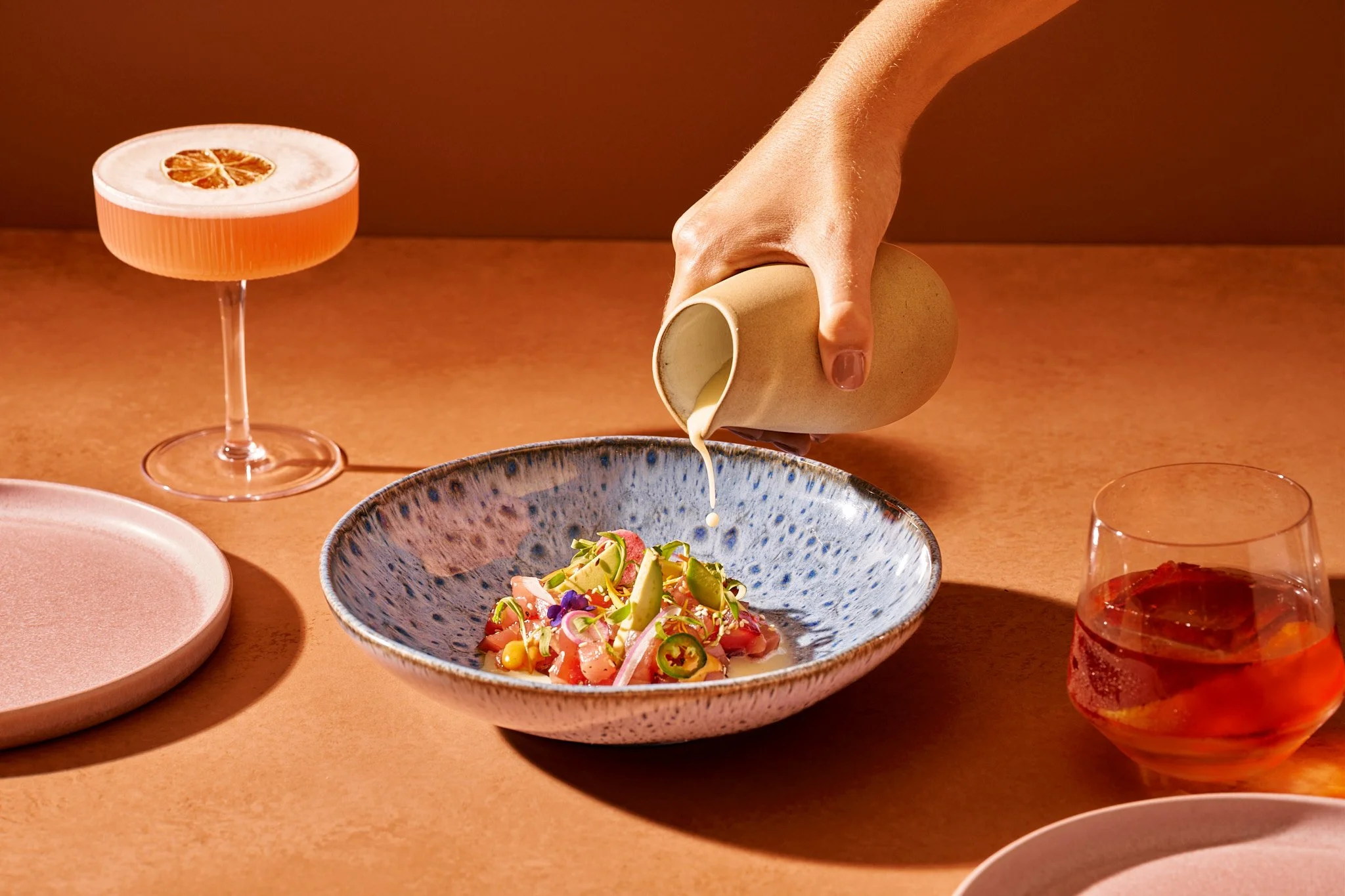 Food Photography for Richard Sandoval Hospitality featuring a person pours a creamy sauce onto a colorful salad in a blue speckled bowl, with pink cocktails and empty plates on a warm-toned table.
