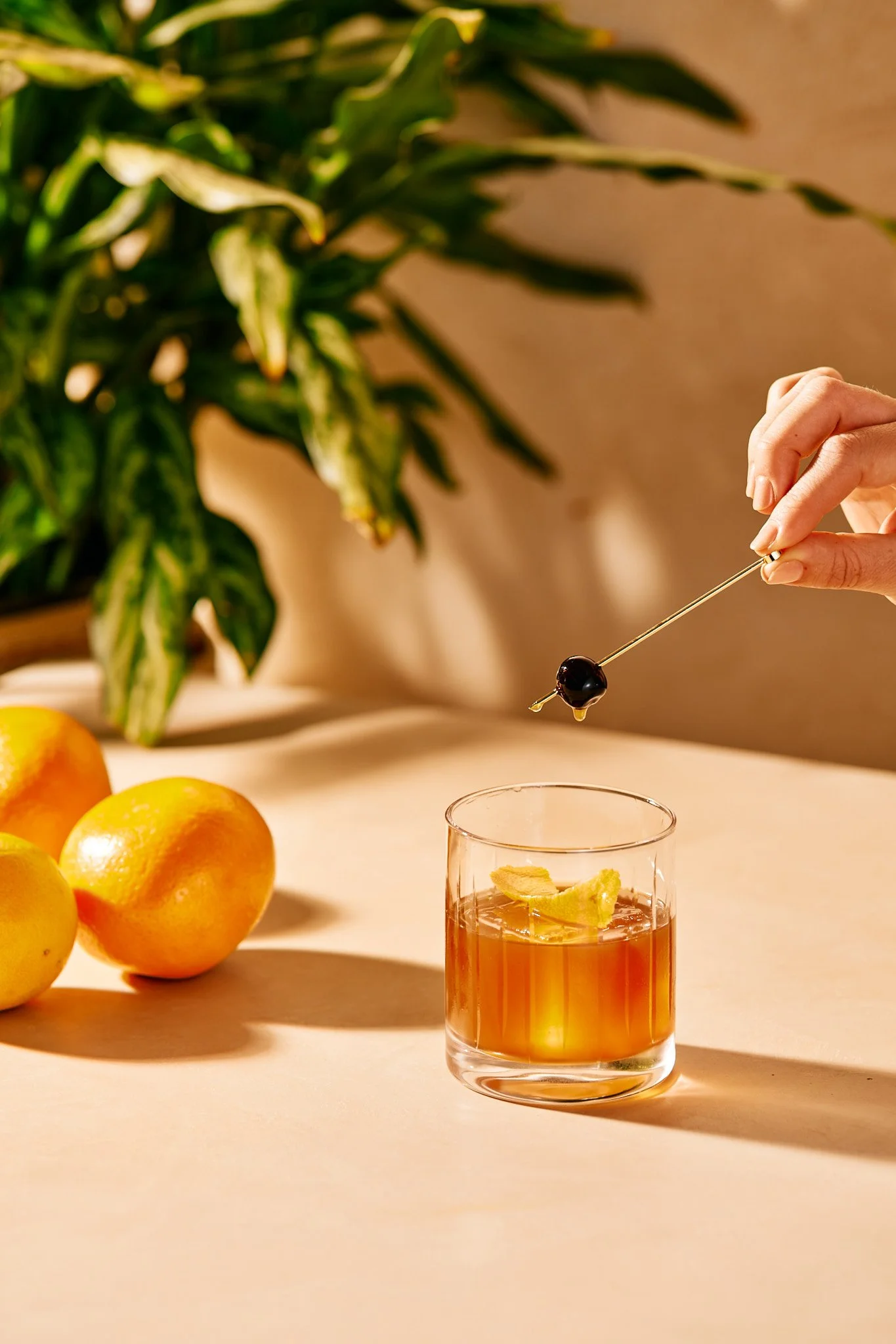 Beverage photography  for Richard Sandoval of hand holding a cocktail pick with a black olive above a glass of an orange-colored drink with lemon slices inside, on a light-colored table with three oranges and a leafy green plant in the background.