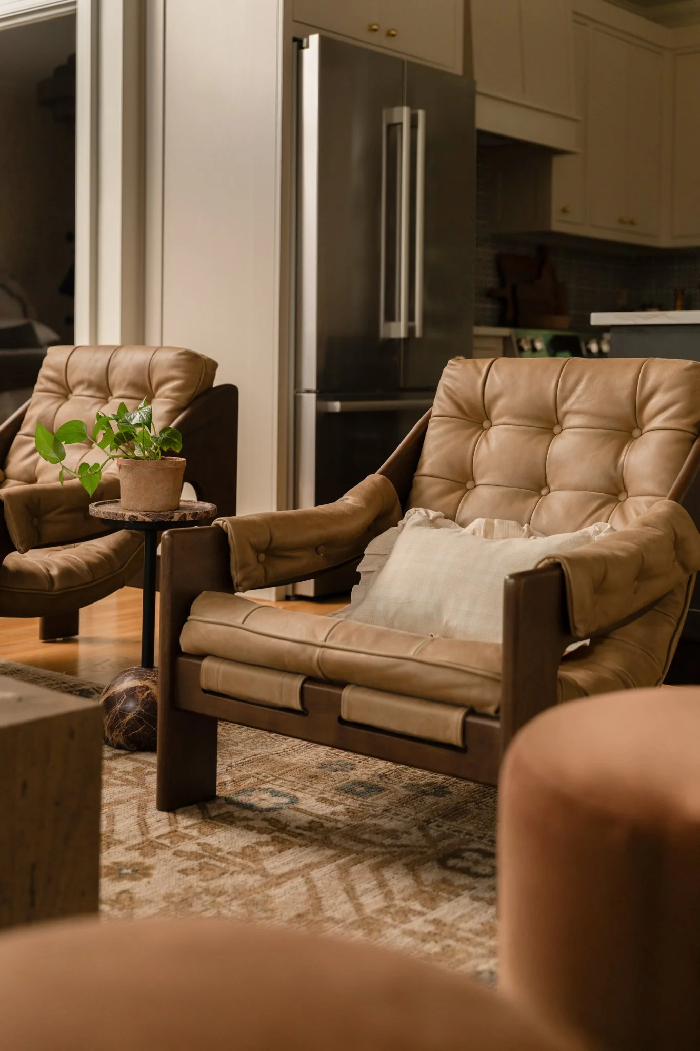Living room with tan leather armchairs, a small side table with a potted plant, and a patterned area rug, with a refrigerator and kitchen cabinets in the background.