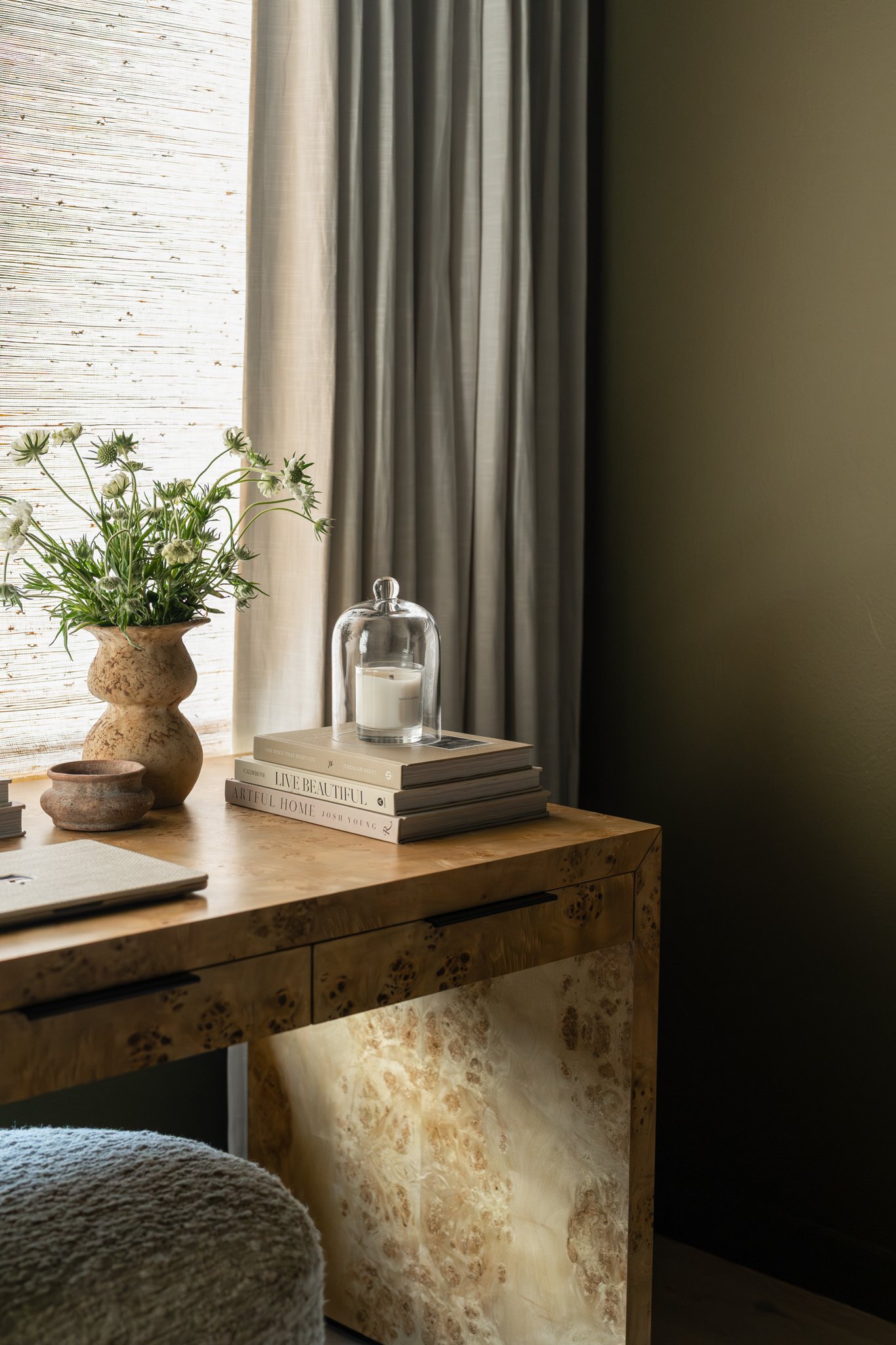 A wooden desk with a stack of books, a glass candle holder, a potted plant, and a small round container, set beside a window with curtains.