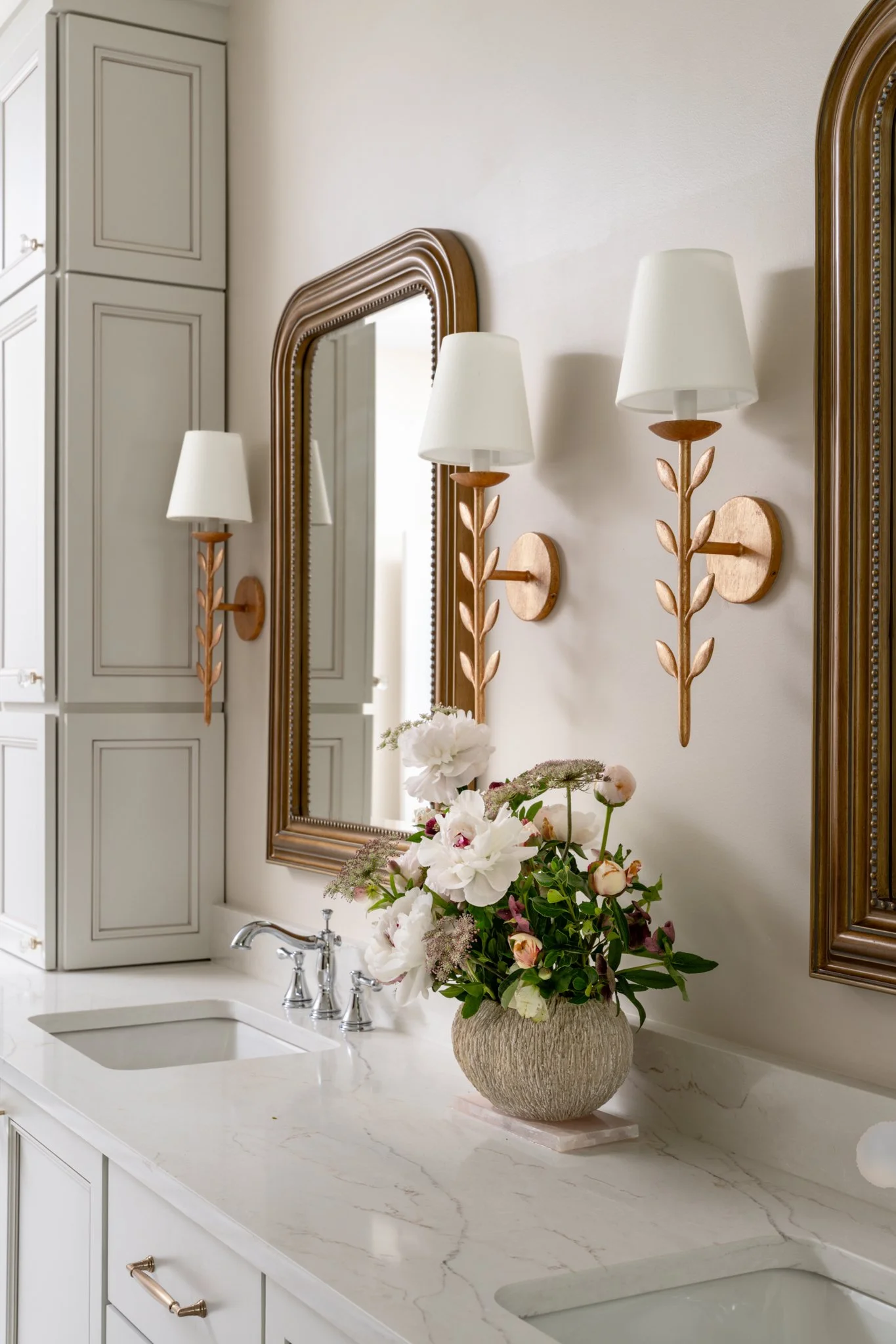 Bathroom with a marble countertop, two sinks, a large mirror, two wall-mounted wooden sconces with white lampshades, a white cabinet on the left, and a vase with white and pink flowers.