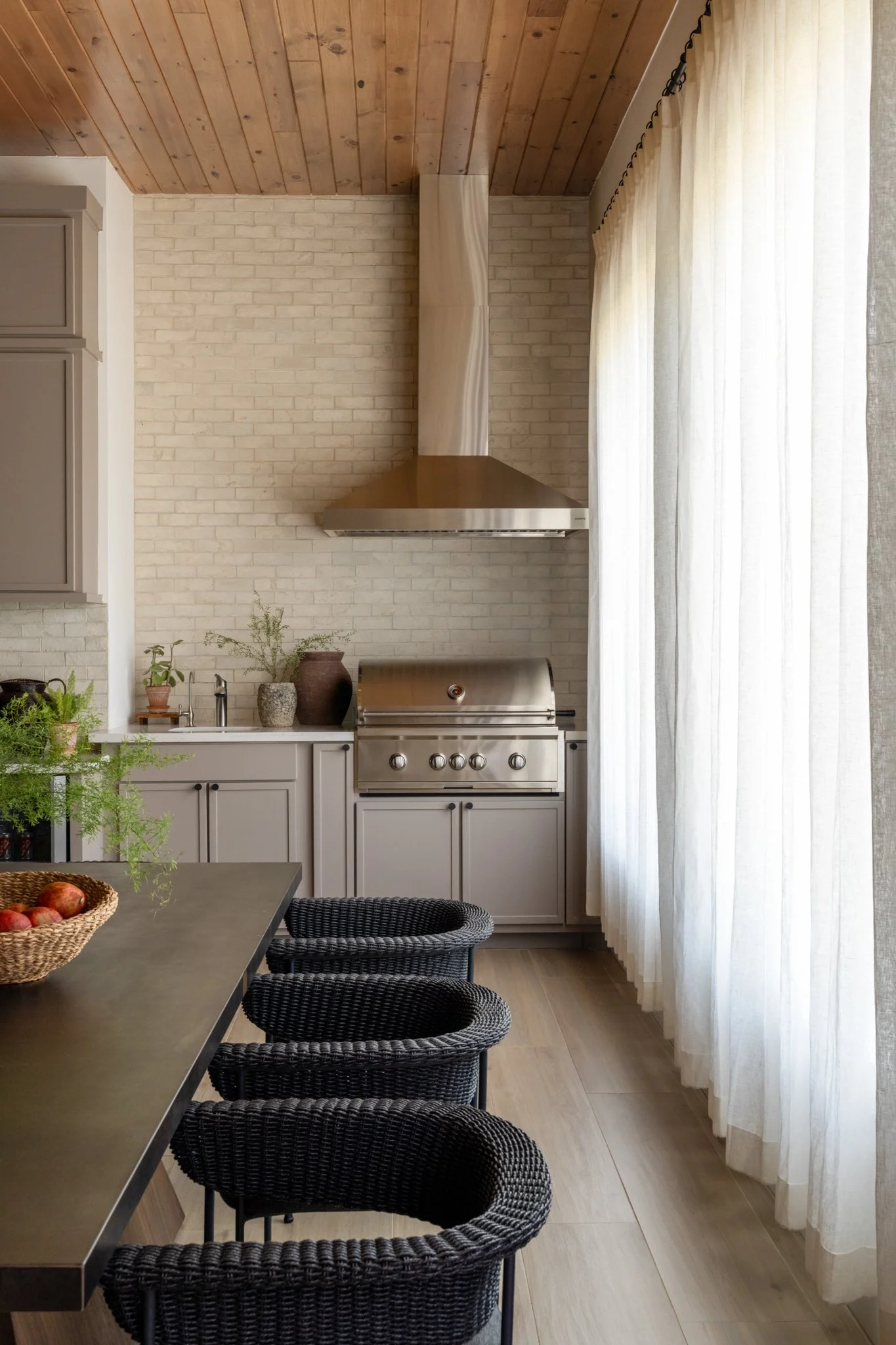 A modern kitchen corner with a stainless steel barbecue grill, a white brick wall, a stainless steel range hood, beige cabinetry, potted plants, and sheer white curtains.