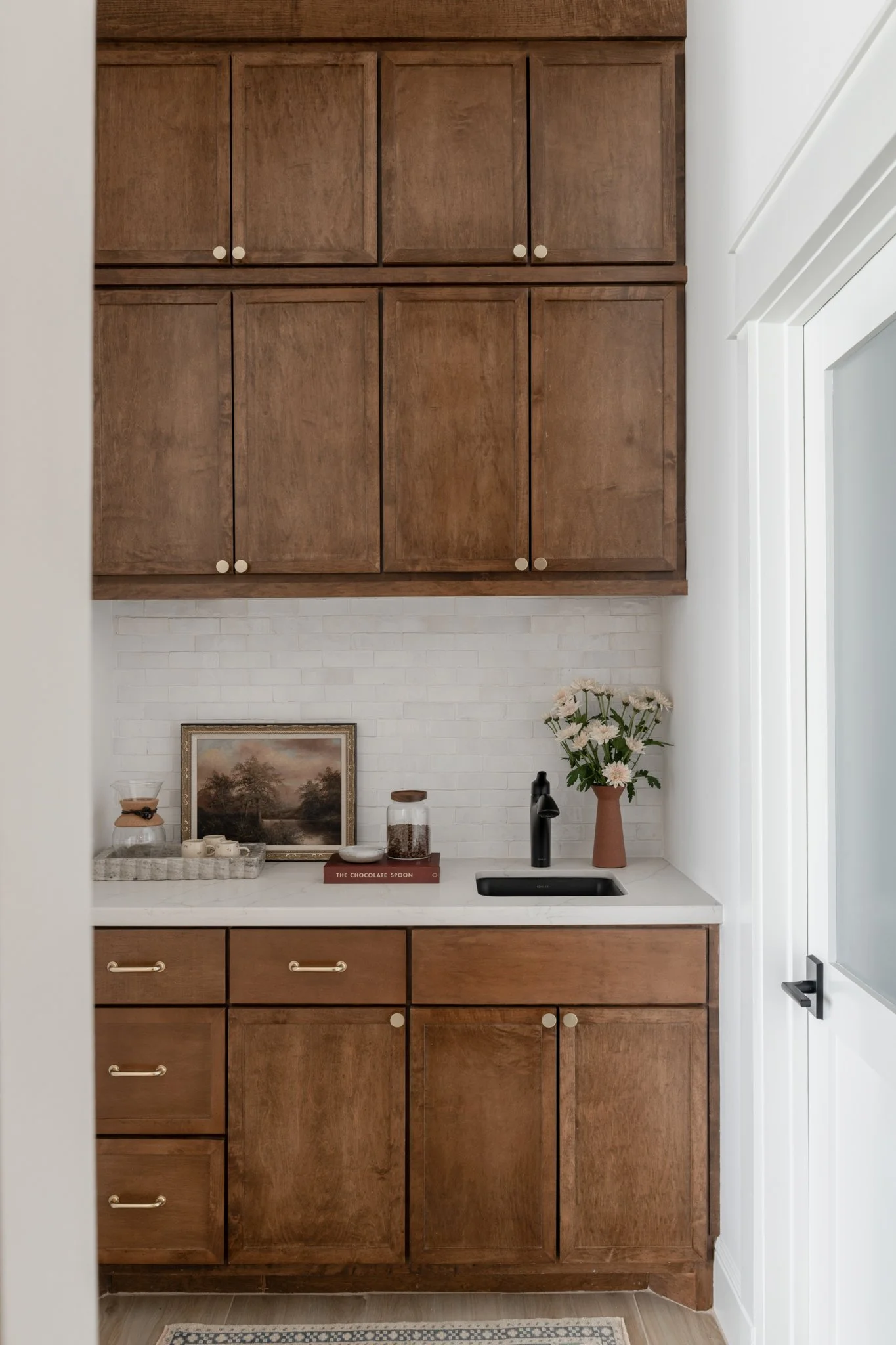 Kitchen corner with wooden cabinets, a white countertop, a black sink with a black faucet, a vase of pink flowers, a framed landscape picture, a glass jar, and a book titled 'The Chocolate Spoon'.