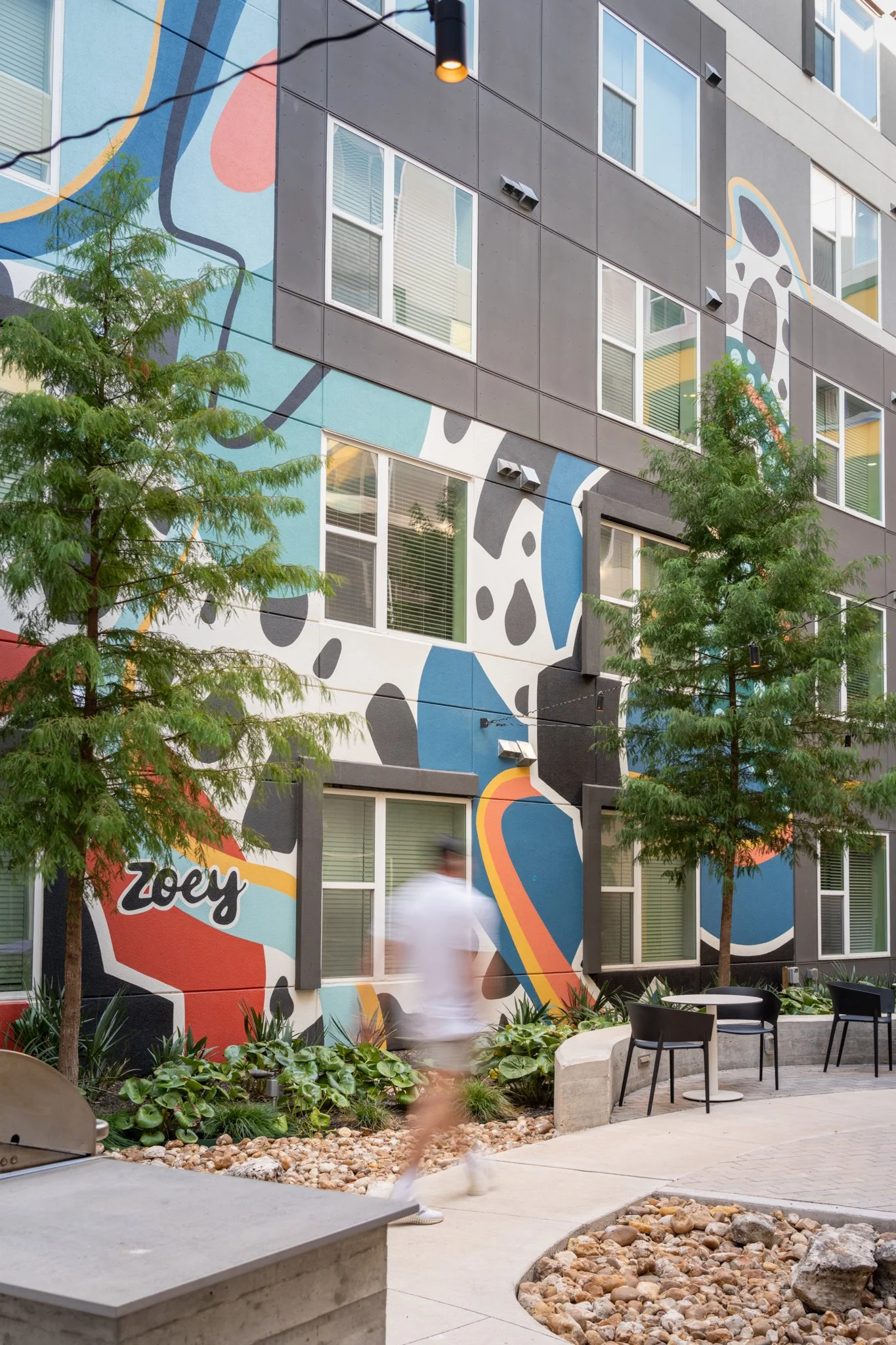 Architectural Photography featuring outdoor patio area of a modern multi-story apartment building with colorful abstract wall art in Austin Texas. There are two green trees, a person walking, and black chairs with small tables in the seating area. 
