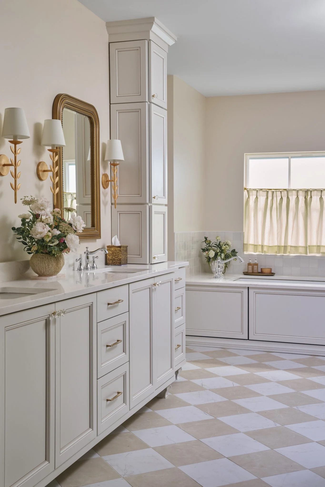 A bright, elegant bathroom with white cabinetry, gold hardware, floral arrangements, a mirror, wall-mounted lights, and a window with a green and beige curtain.