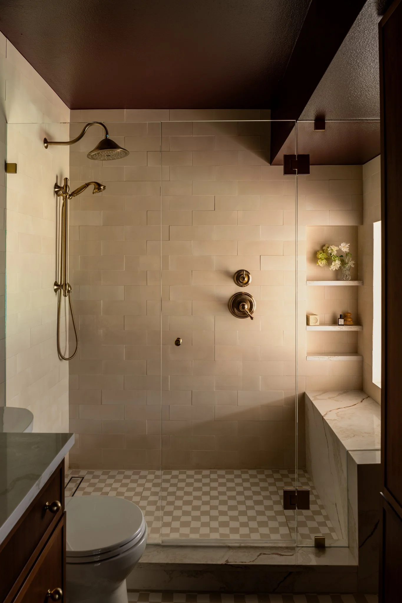 Modern bathroom shower with brass fixtures, a glass door, built-in shelves with decorative items, and a window with natural light.