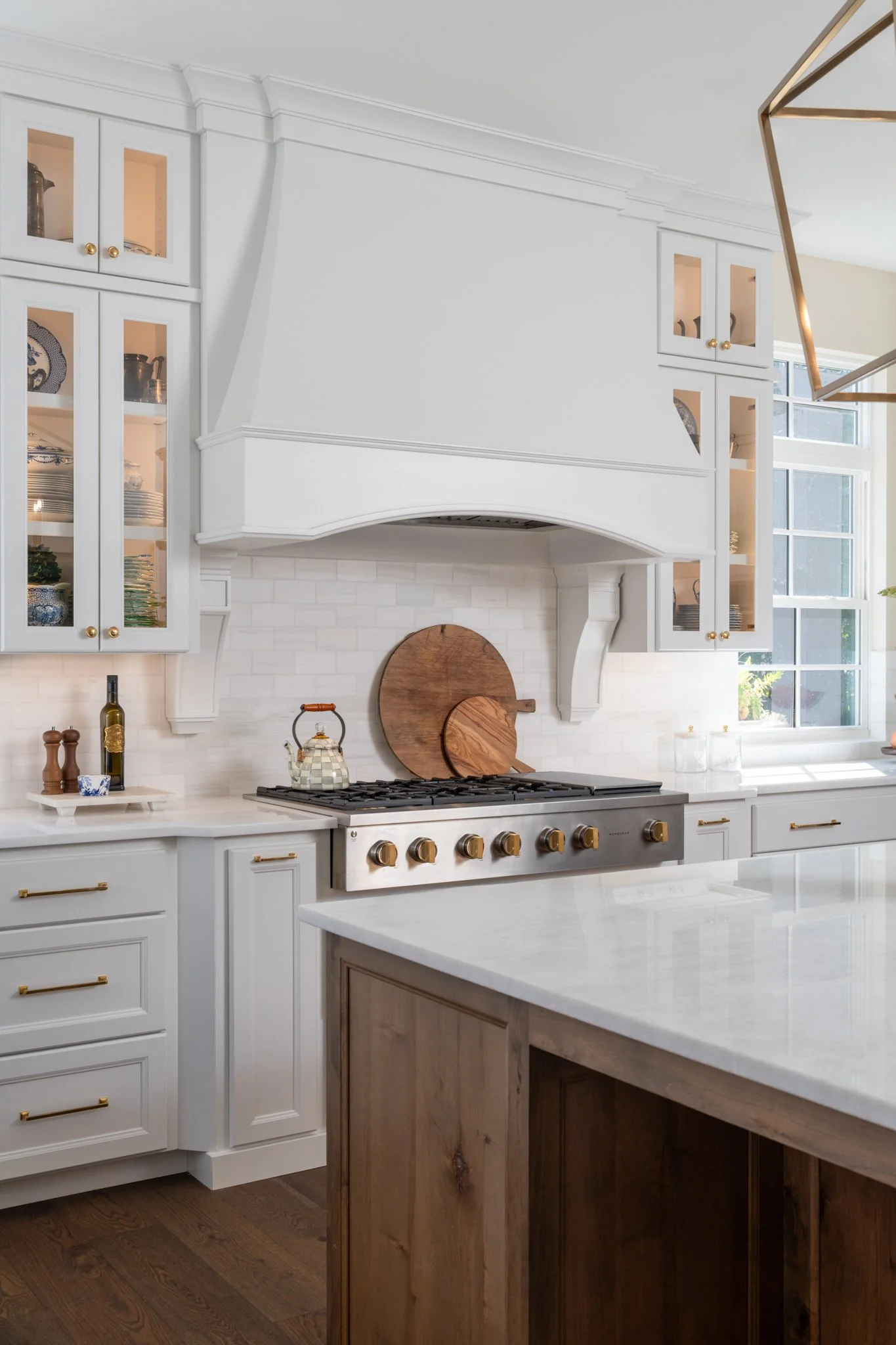 White kitchen with built-in cabinets, a stove with brass knobs, a white countertop, and a kitchen island with a wood base. There are decorative items and a window letting in natural light.