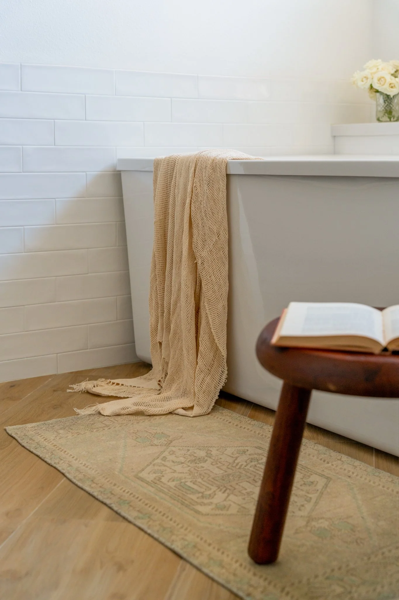 A cozy bathroom corner with a white bathtub, a beige blanket draped over the edge, a small wooden stool with an open book, a patterned rug on wooden flooring, and a vase of white roses on the ledge.