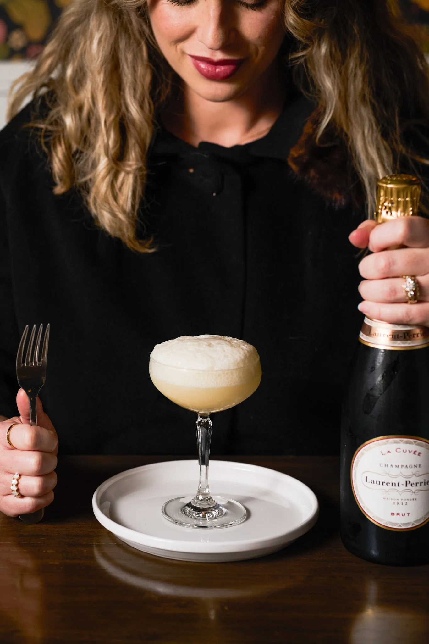 Beverage Photography at Blush Restaurant in San Antonio, Texas with woman holding a fork in one hand and a bottle of champagne in the other, with a coupe glass of frothy cocktail on a white plate in front of her, on a wooden table.