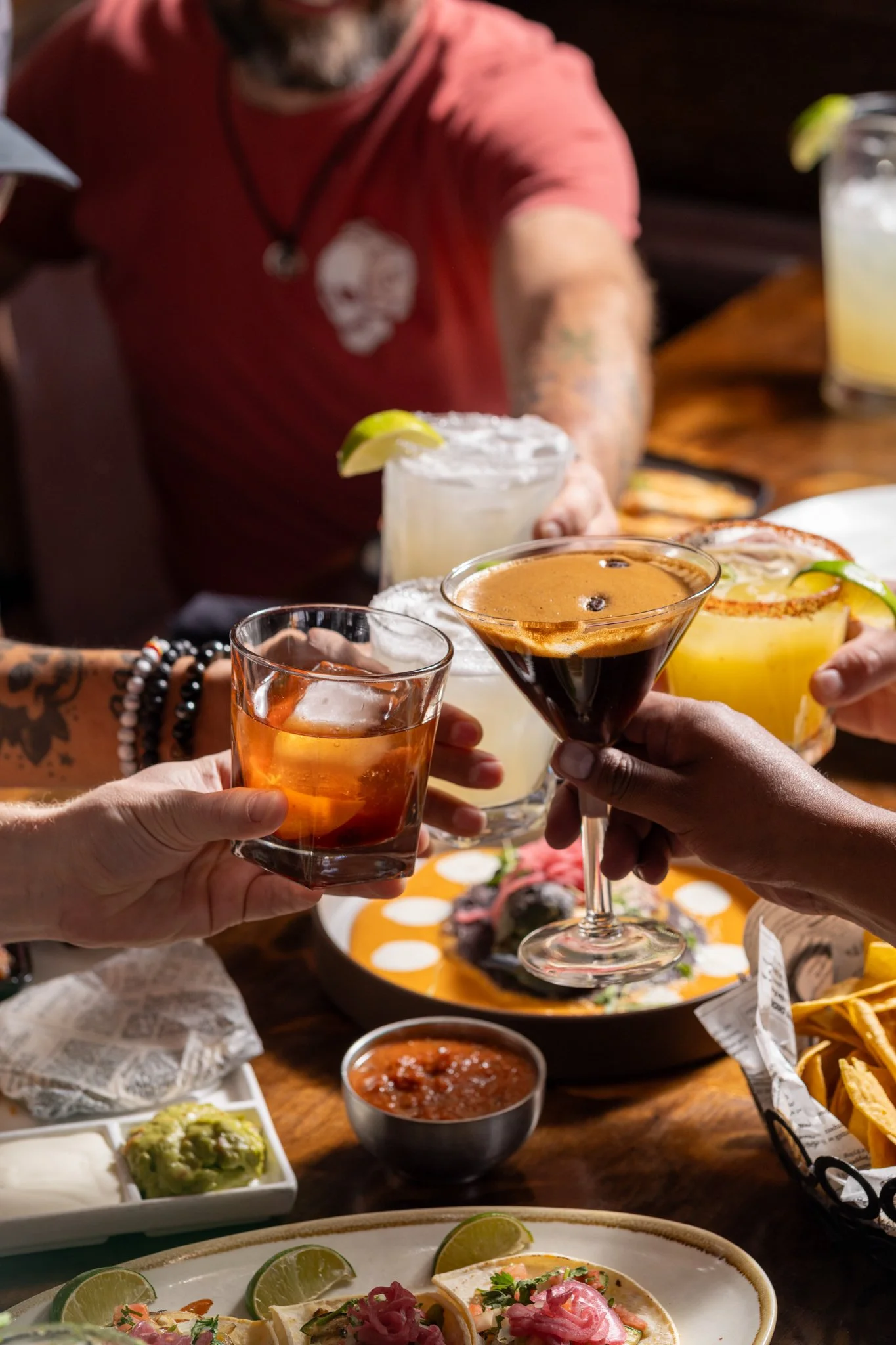 Food photography for Richard Sandoval restaurant Venga Venga in Aspen, CO People clinking glasses of cocktails and beer at a festive meal, with plates of food, guacamole, salsa, and tacos on the table.