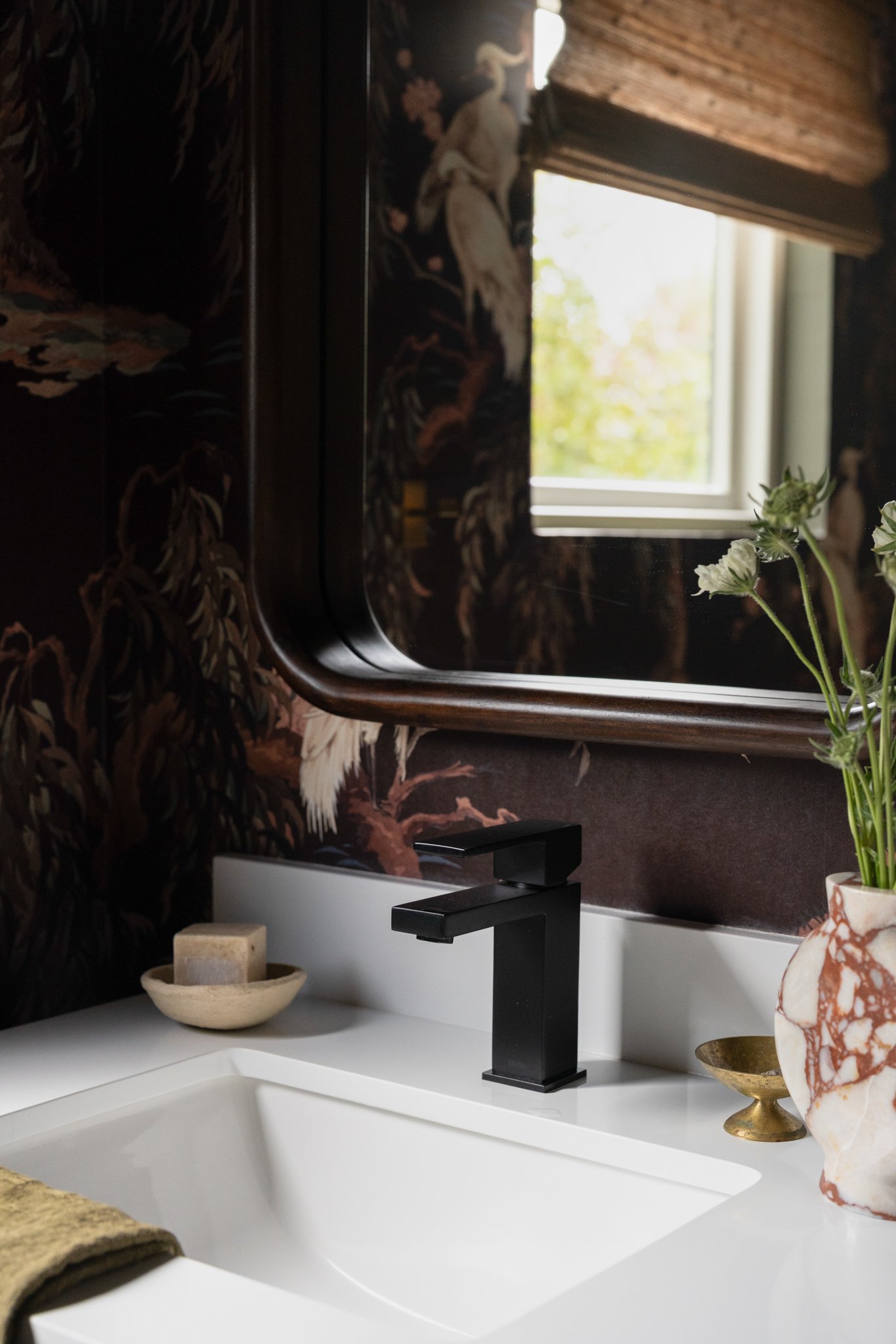 Close-up of a bathroom sink with a black faucet, a beige soap dish, and a gold dish with a decorative item on a white countertop. A large mirror reflects a window with greenery outside, and a vase with white flowers is placed beside the sink.