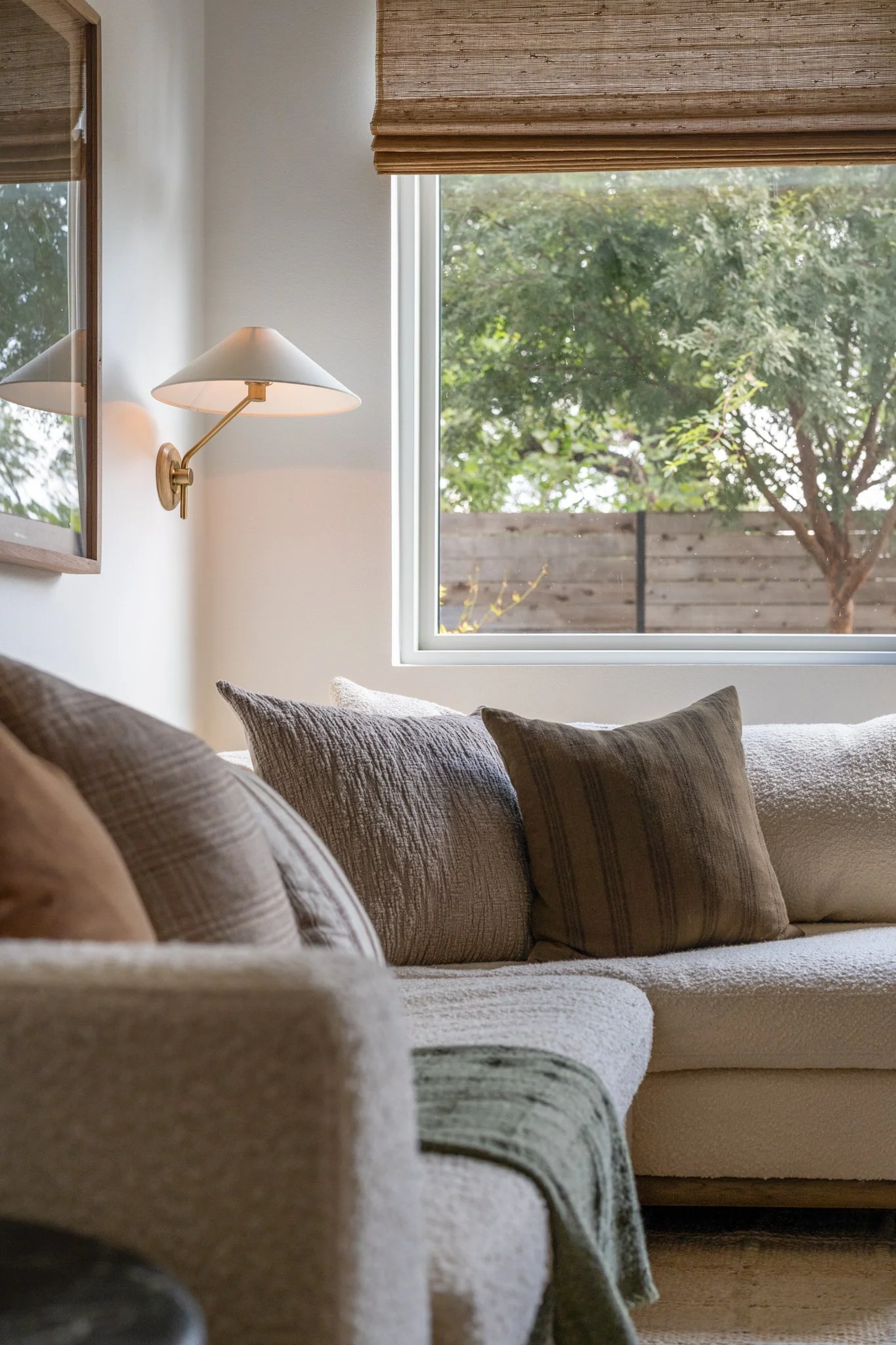 Living room with beige sofa, brown and gray throw pillows, a wall-mounted lamp, and a large window showing green trees outside.