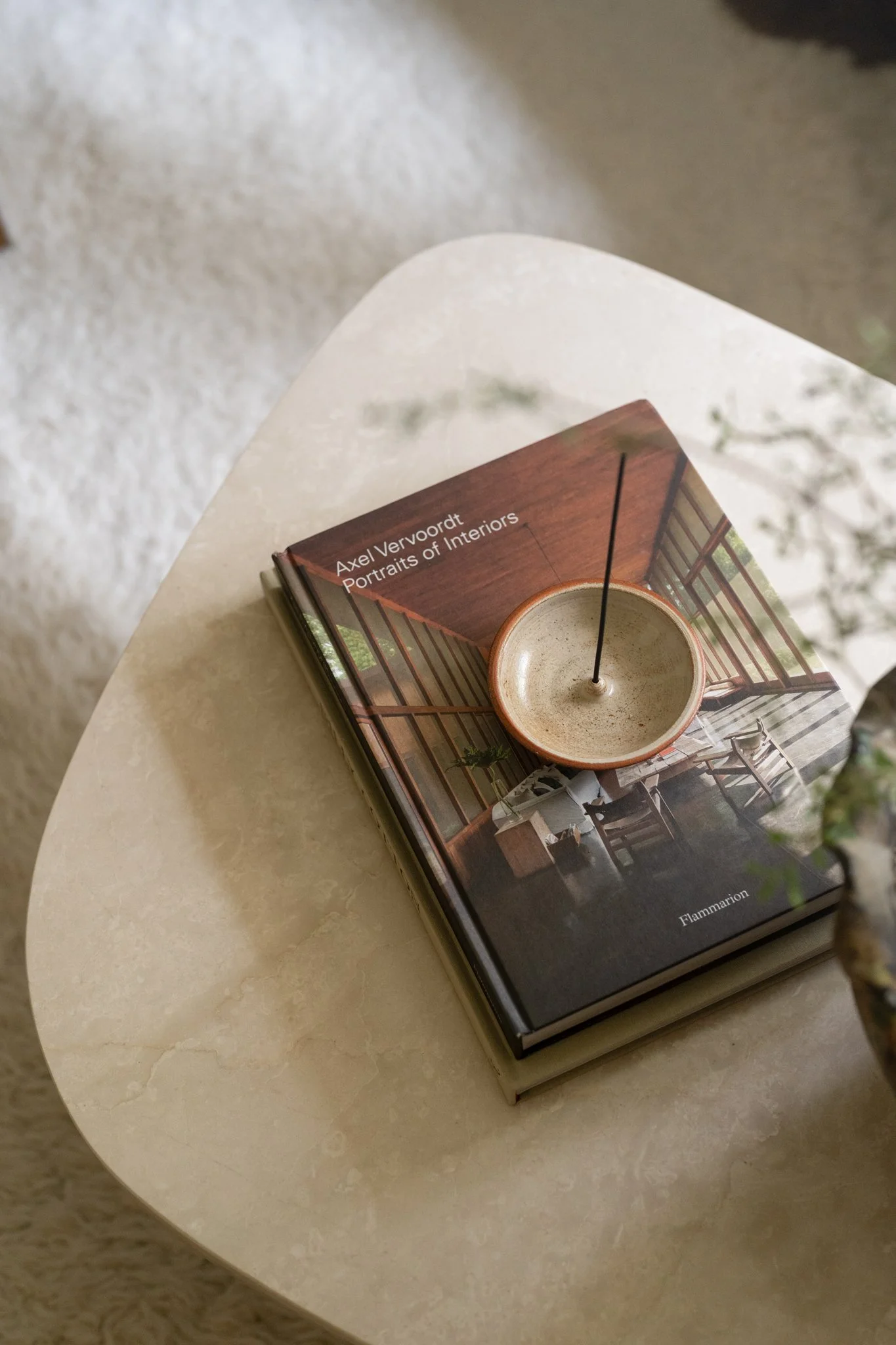 A hardcover book titled 'Portraits of Interiors' by Axel Vervoort lies on a light-colored table with a small decorative bowl on top of it. The bowl contains a thin black reed.