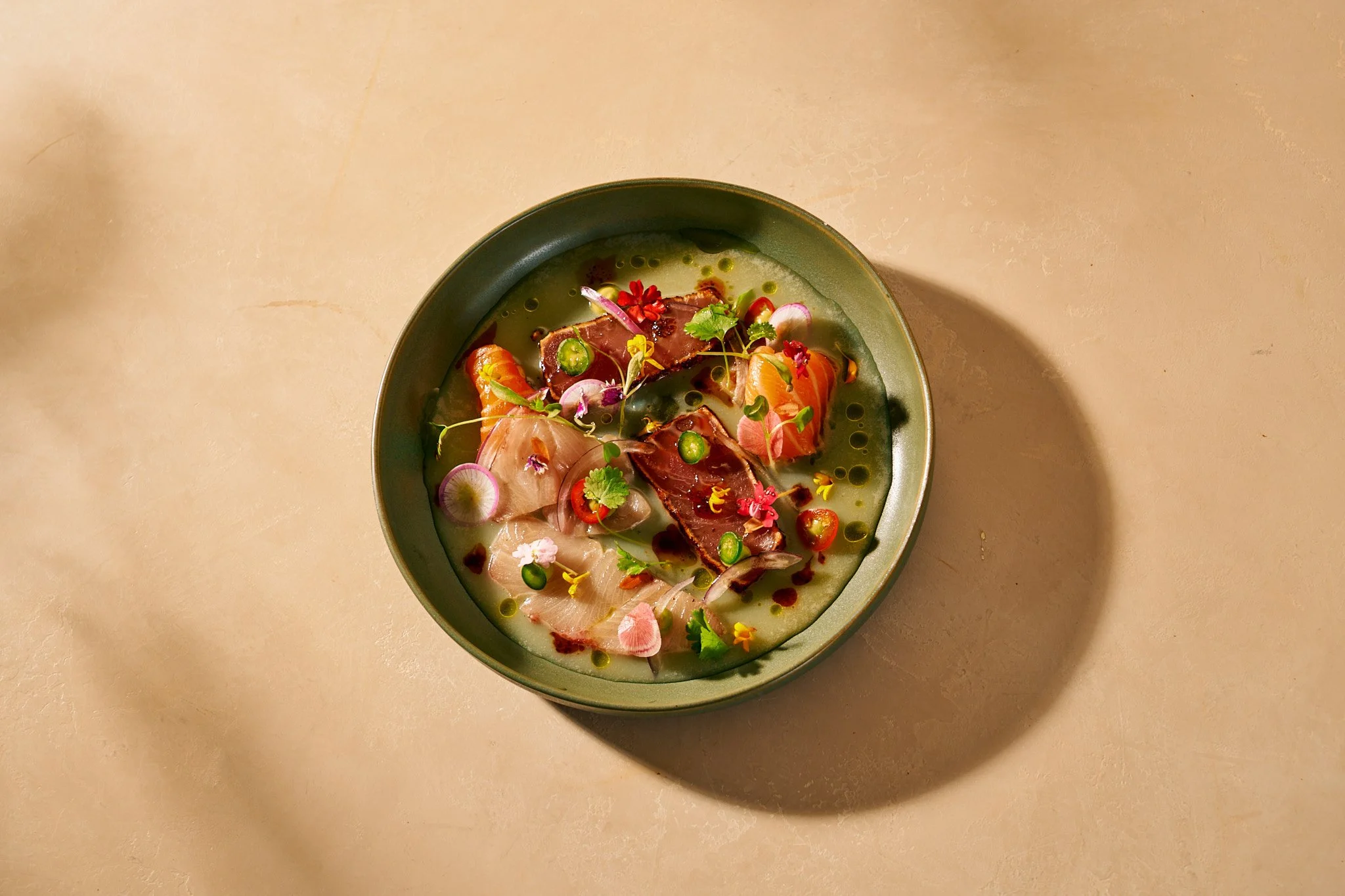 Beverage photography  for Richard Sandoval Hospitality featuring  A bowl of sashimi with various slices of raw fish, garnished with microgreens, edible flowers, and thinly sliced radishes, served with a green liquid sauce.