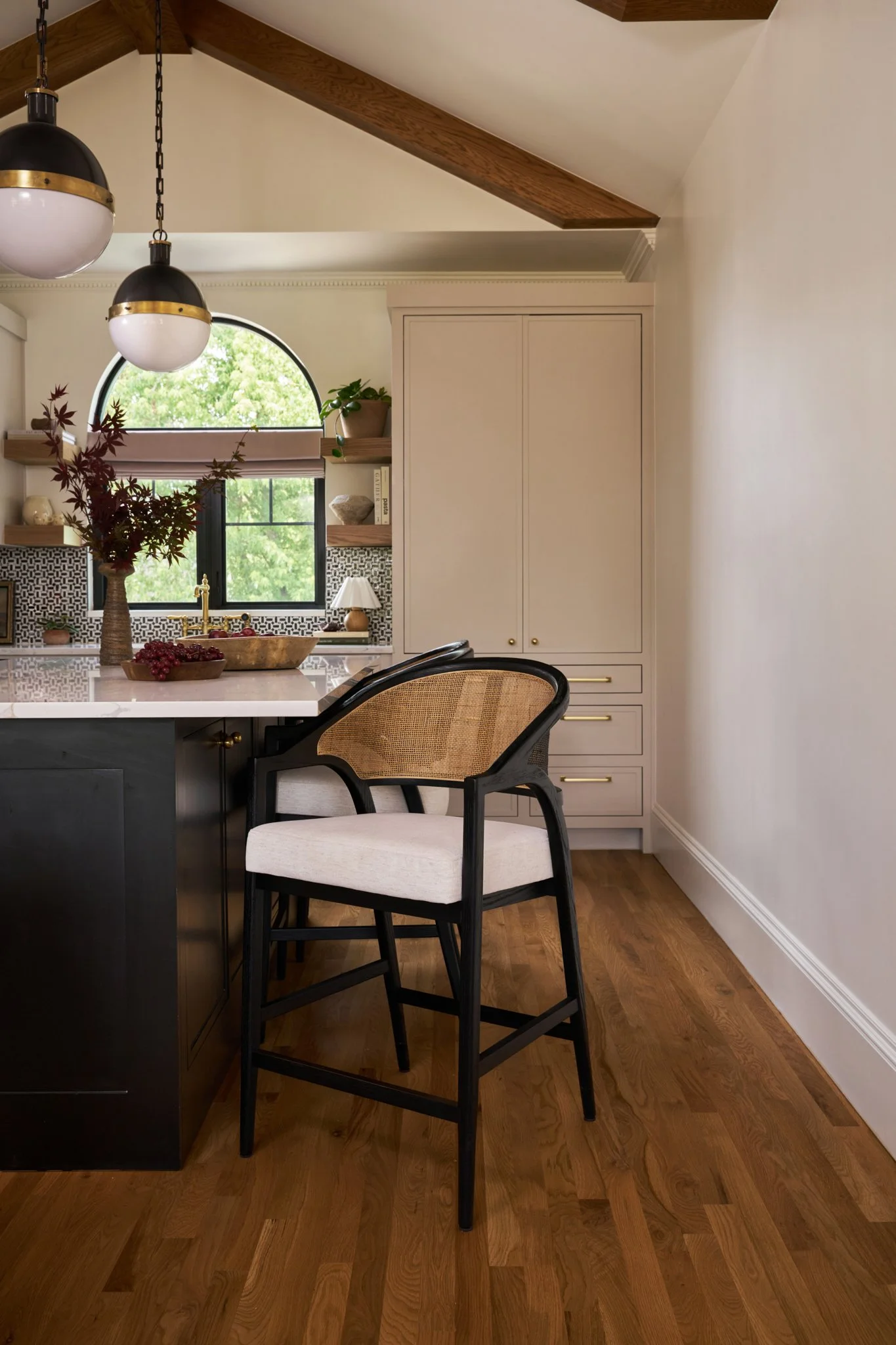 Modern kitchen with a black and beige chair at a white kitchen island, a window with greenery outside, floating shelves with decor, and hardwood flooring.