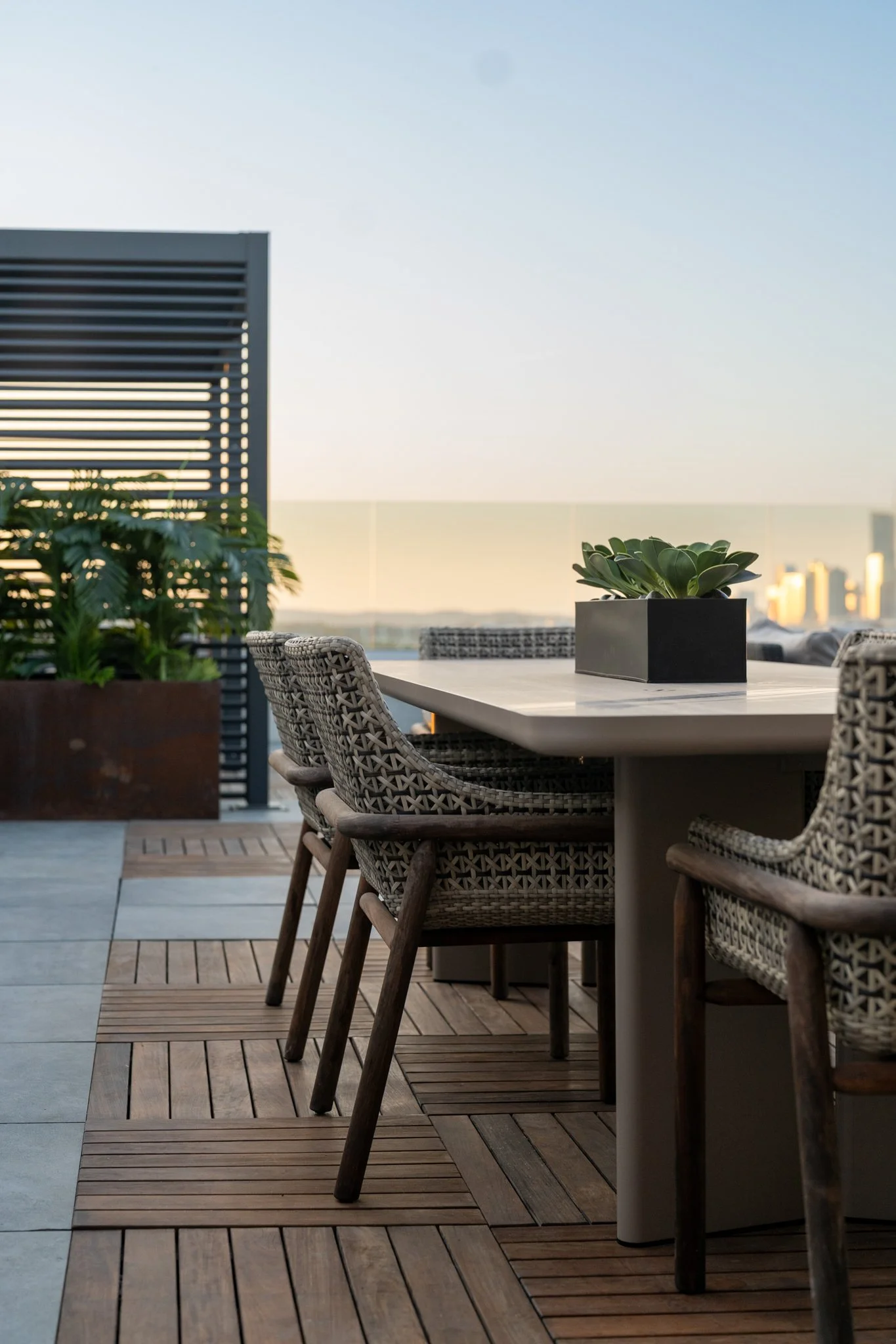 Rooftop Patio of Zoey Apartments in Austin Texas featuring outdoor patio with a rectangular table, woven chairs, potted plants, and city skyline in the background during sunset.