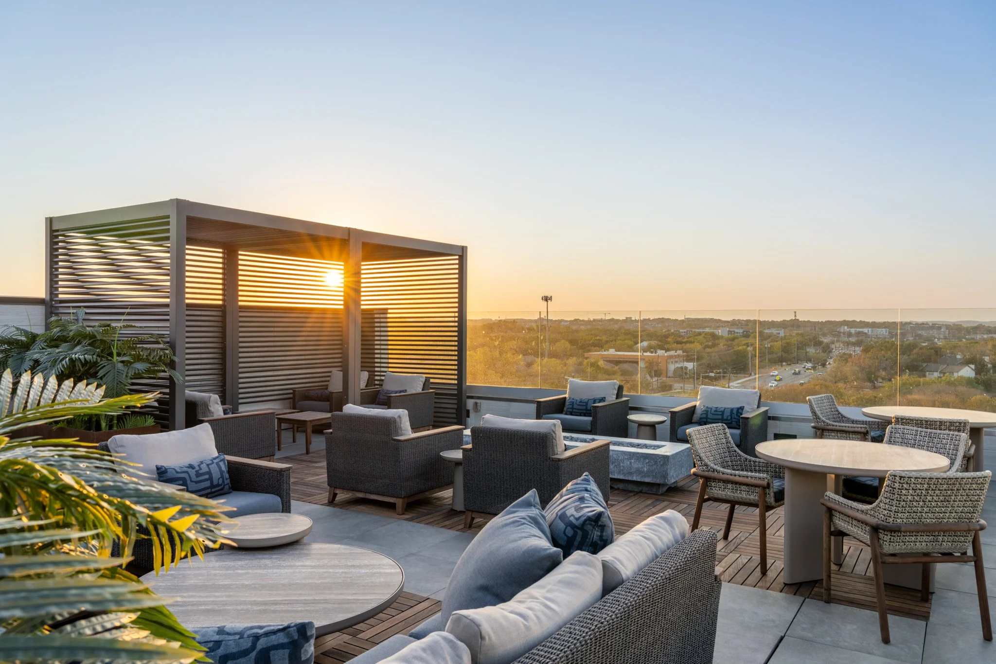 Architectural Photography featuring rooftop Patio of Zoey Apartments in Austin Texas. Rooftop lounge area with outdoor seating, including cushioned chairs and tables, with a wooden deck and a privacy canopy, overlooking a cityscape at sunset.