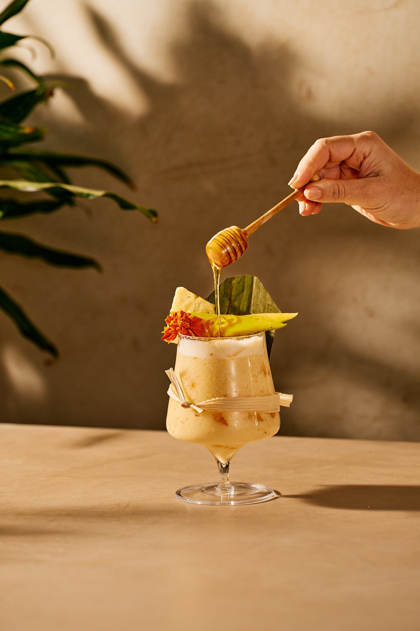 Beverage photography  for Richard Sandoval Hospitality featuring  A tropical cocktail garnished with pineapple, orange flower, and a leaf, with honey being drizzled into it from a honey dipper.