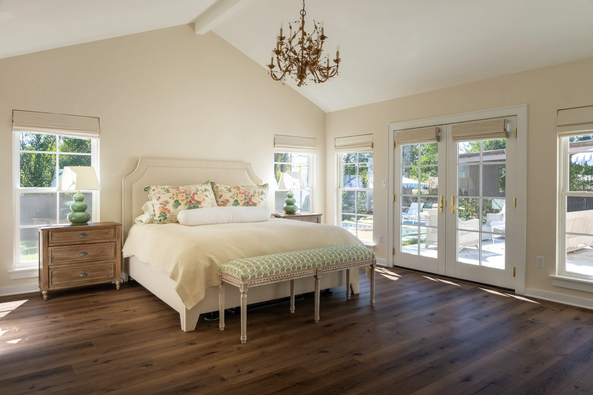 Bright bedroom with a large bed, floral pillows, two wooden nightstands with green lamps, a bench at the foot of the bed, hardwood floors, and large windows and glass doors leading to an outdoor patio.