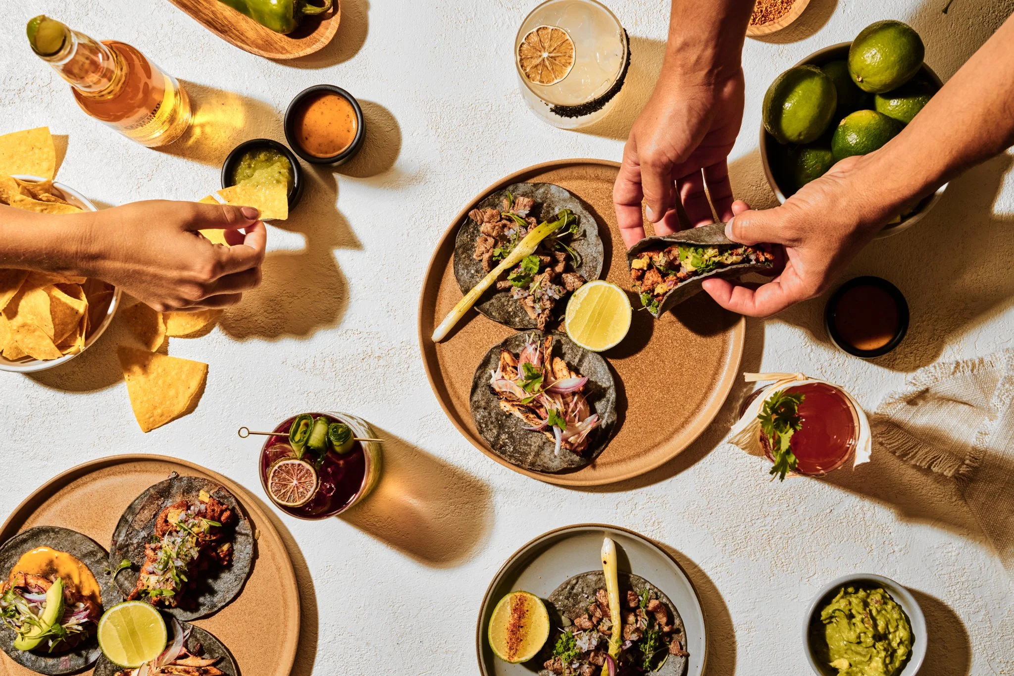 Beverage photography  for Richard Sandoval Hospitality featuring  Top-down view of a Mexican meal featuring tacos, chips, guacamole, lime wedges, and drinks on a white textured surface.