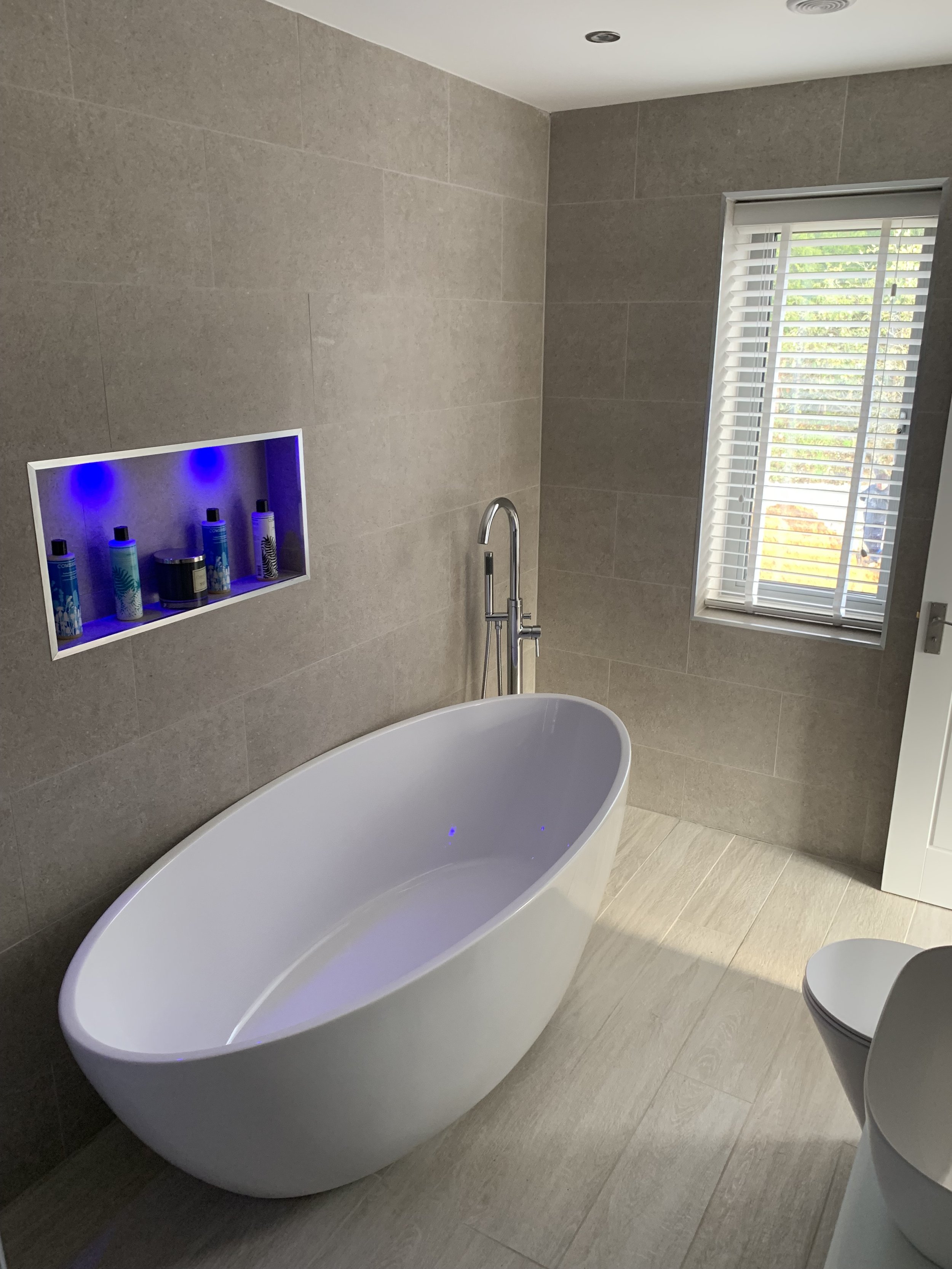 Modern bathroom with a white freestanding bathtub, gray tiled walls, a window with white blinds, and a built-in shelf illuminated by blue lights.