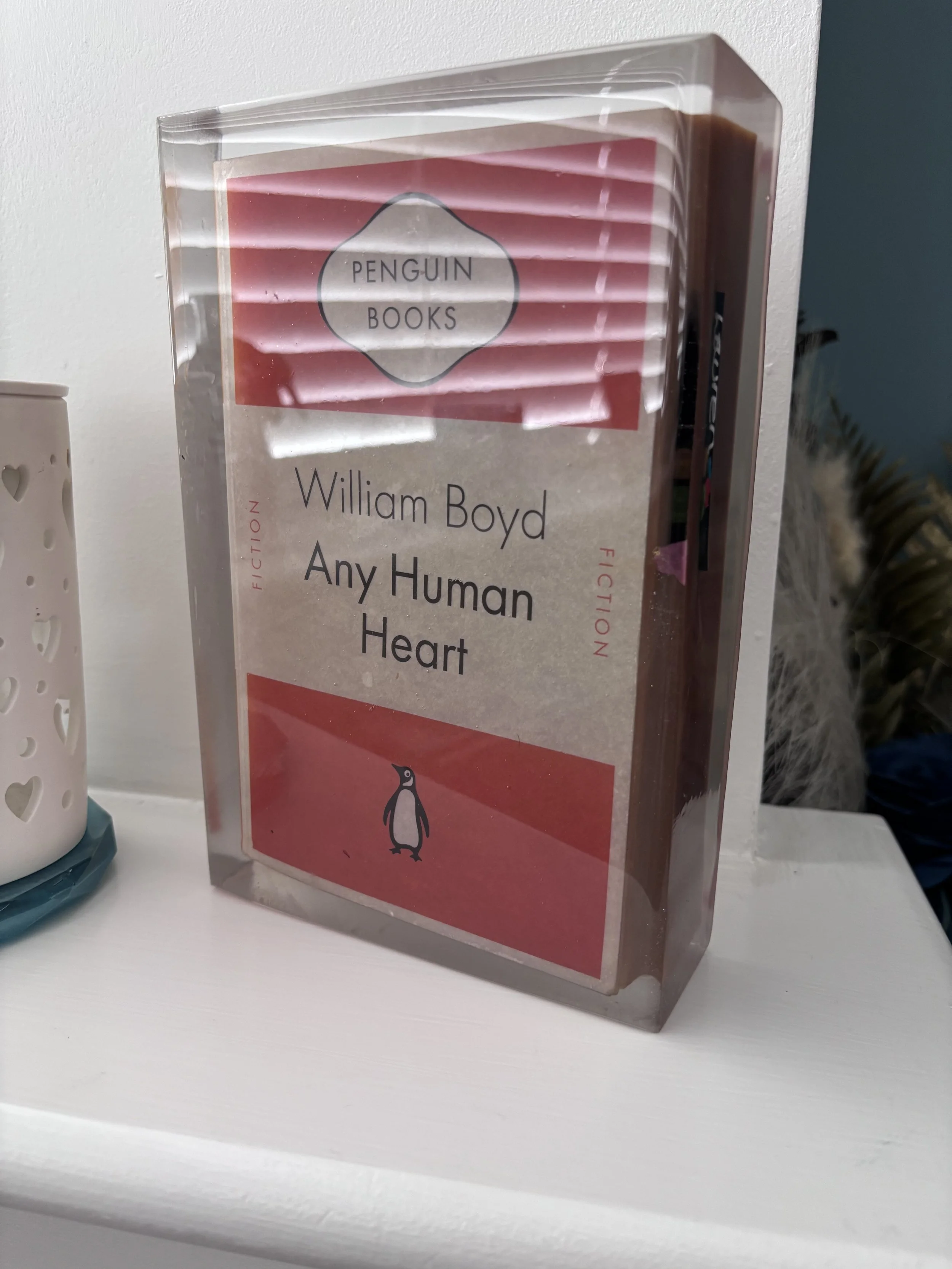 A paperback book titled 'Any Human Heart' by William Boyd, encased in a clear plastic protective cover, positioned on a white surface next to a white decorative item with heart-shaped cutouts.