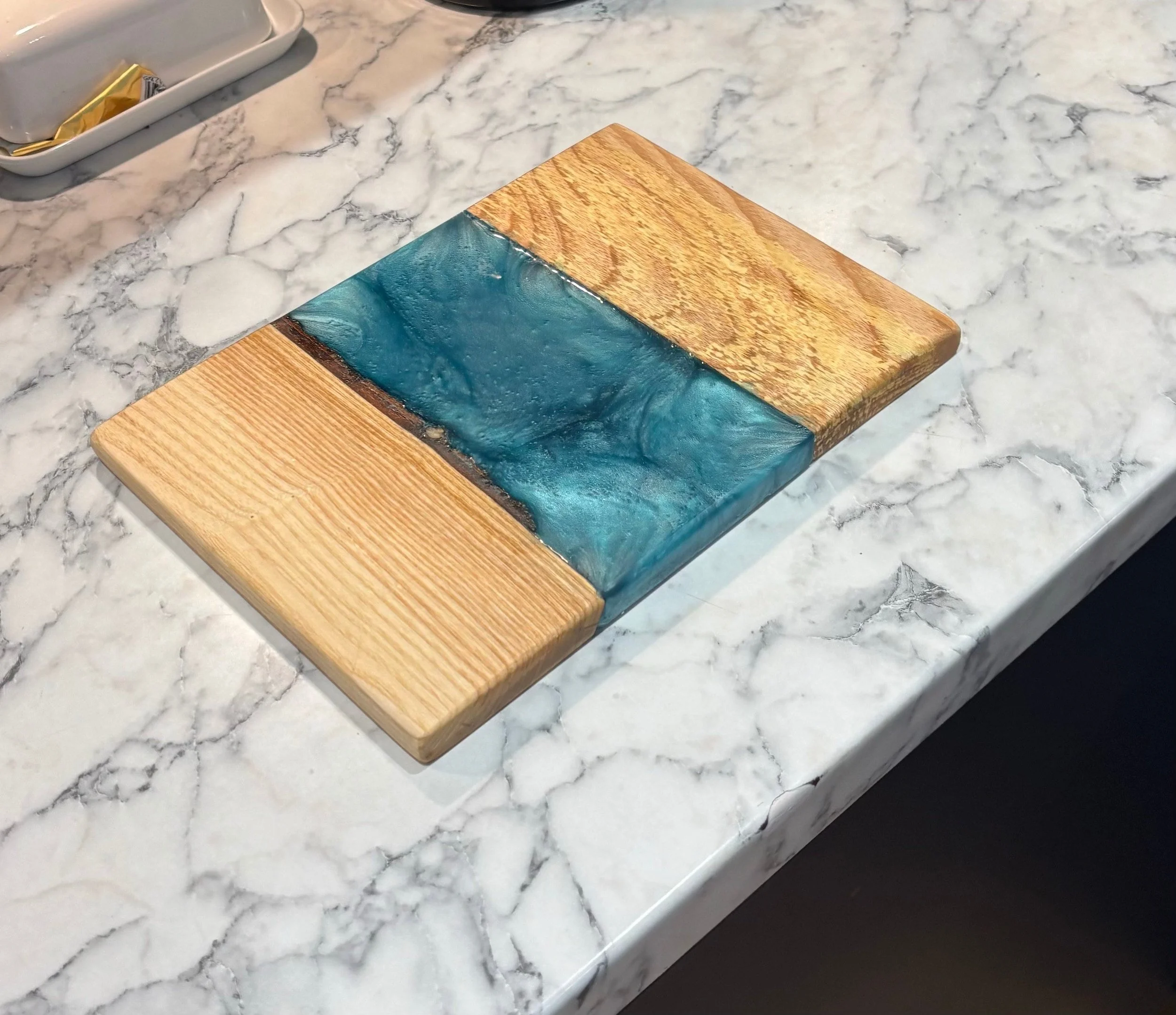 A square wooden and resin art piece placed on a marble surface, featuring natural wood grains and a blue resin design resembling water.