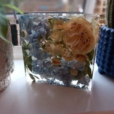 Square clear resin block with preserved blue and cream flowers inside, displayed on a windowsill.