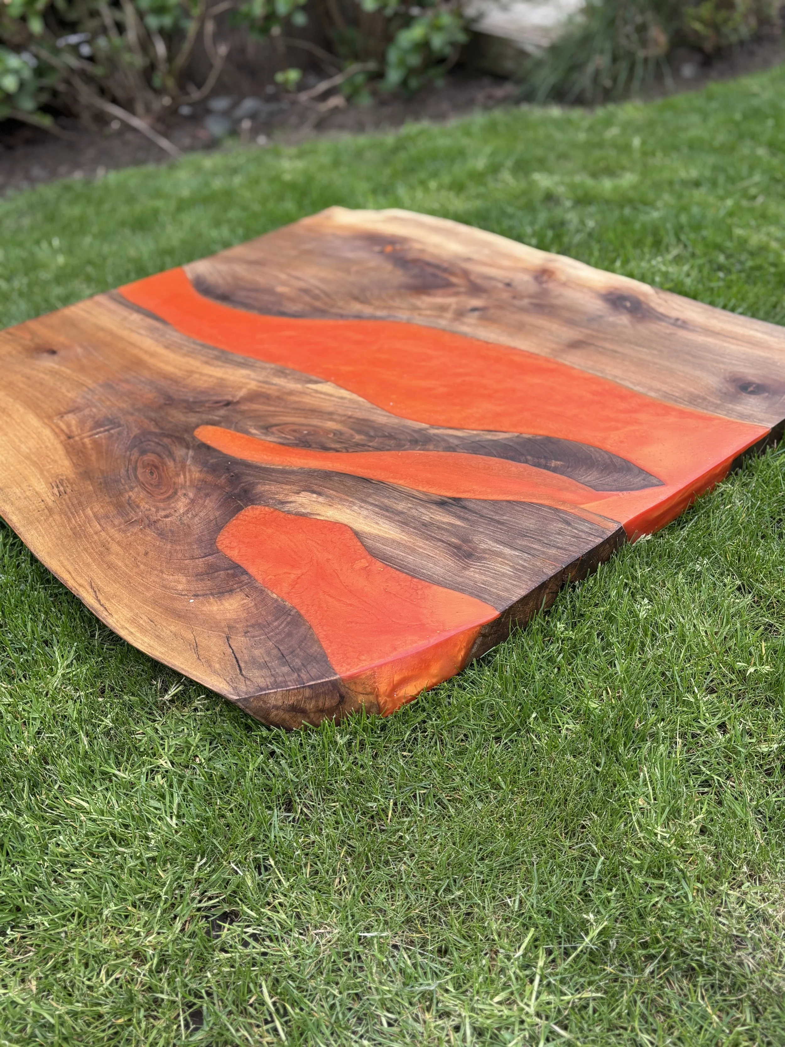 Wooden table with orange epoxy resin accents placed on green grass, with bushes and soil in the background.