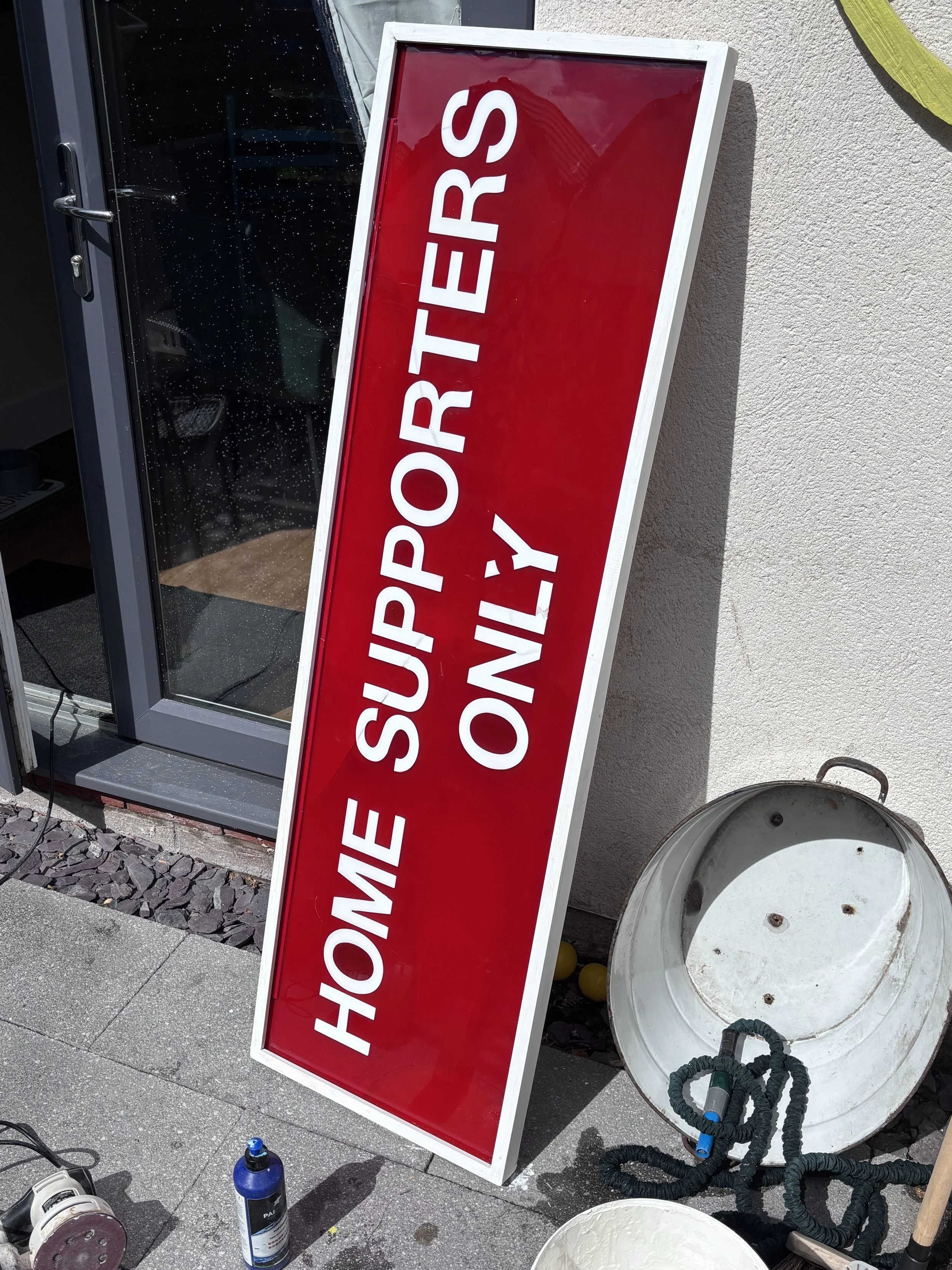 Red rectangular sign with white letters reading "HOME SUPPORTERS ONLY" leaning against a wall outside a building near a glass door, with construction or cleaning tools nearby.