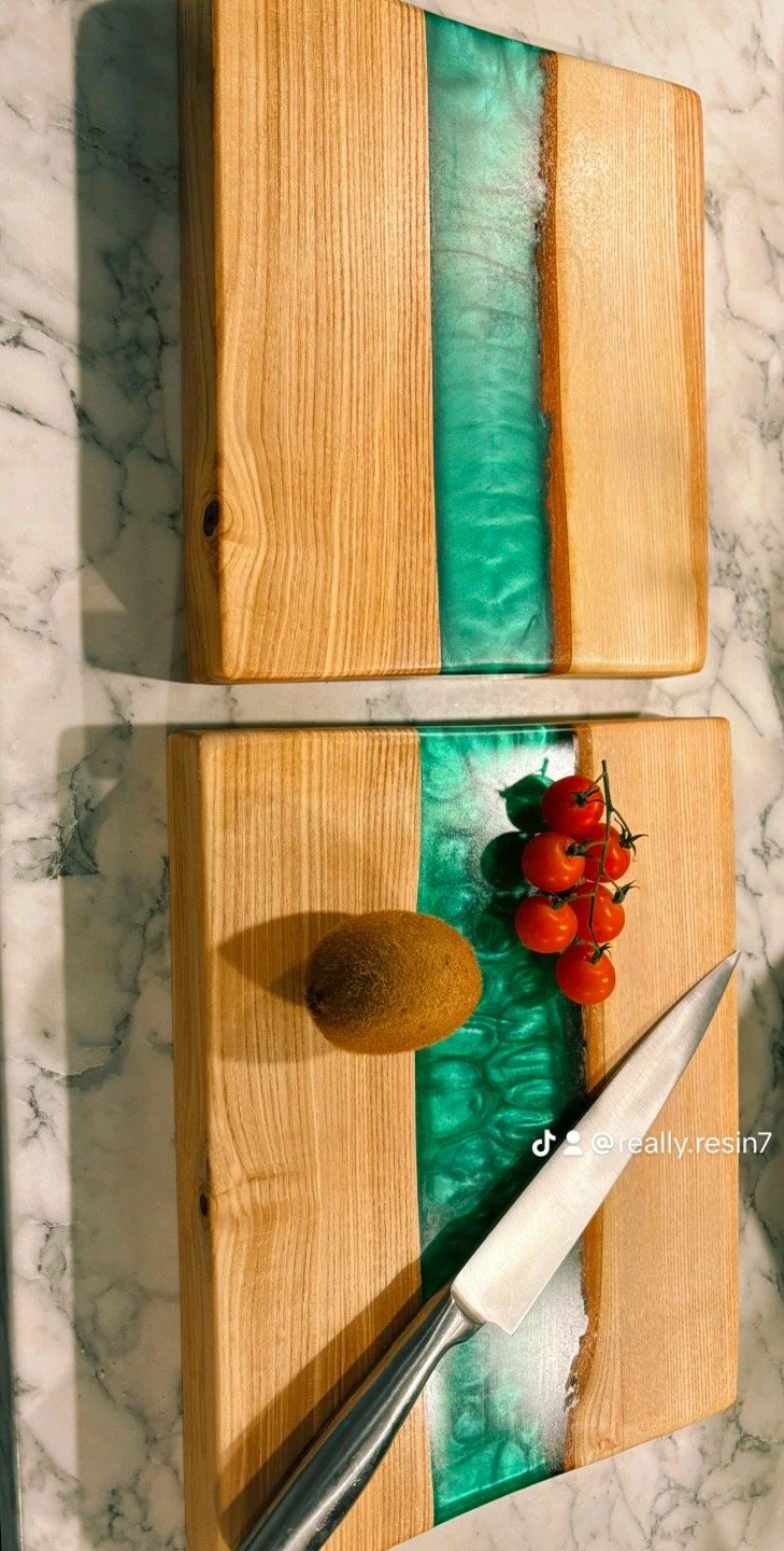 Two wooden cutting boards with turquoise resin inlays, placed on a marble countertop. The bottom board has a kiwi, a bunch of cherry tomatoes, and a chef's knife resting on it.