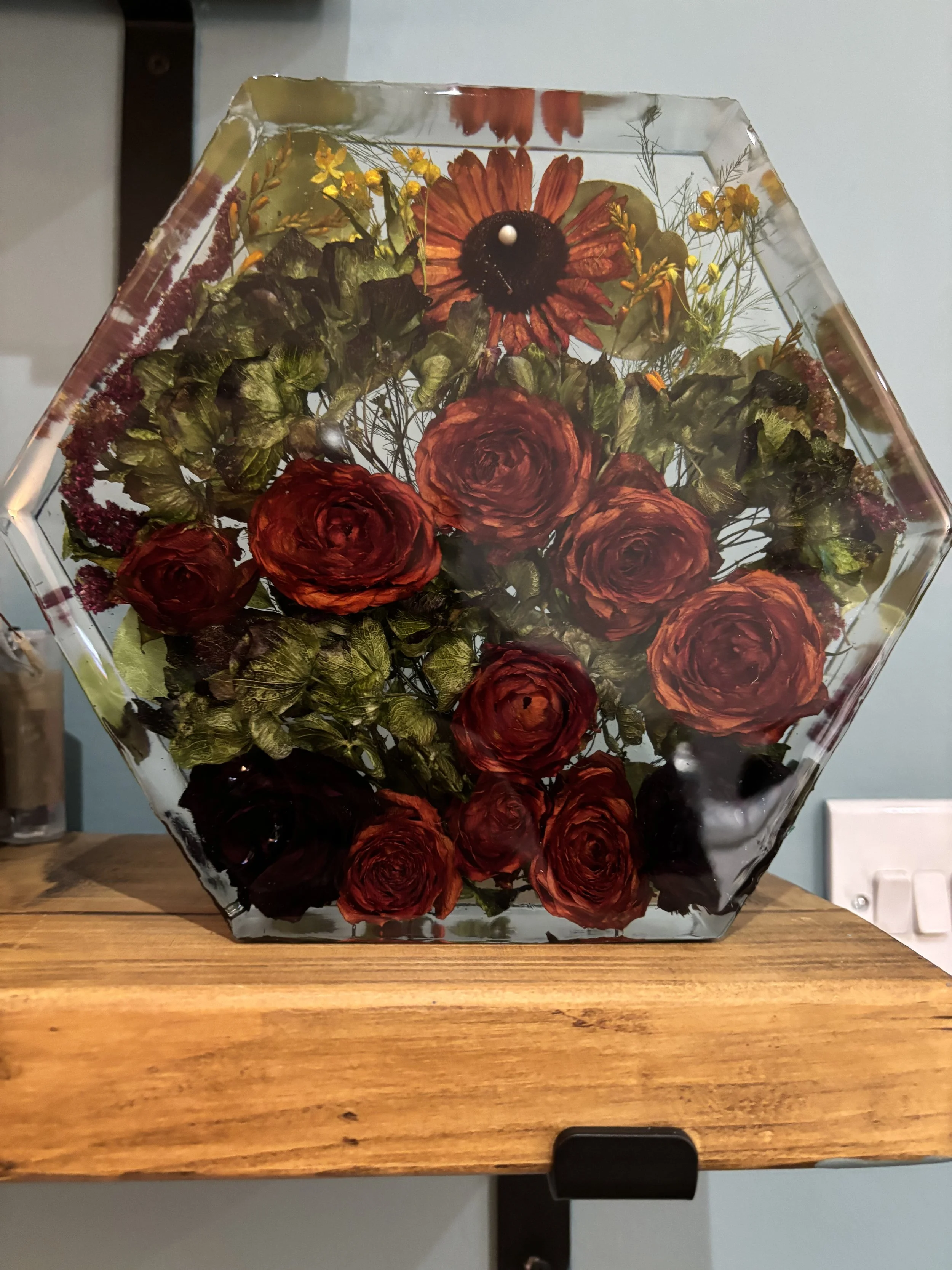 A transparent hexagonal vase with dried red roses, purple and yellow flowers, and green foliage, placed on a wooden surface.