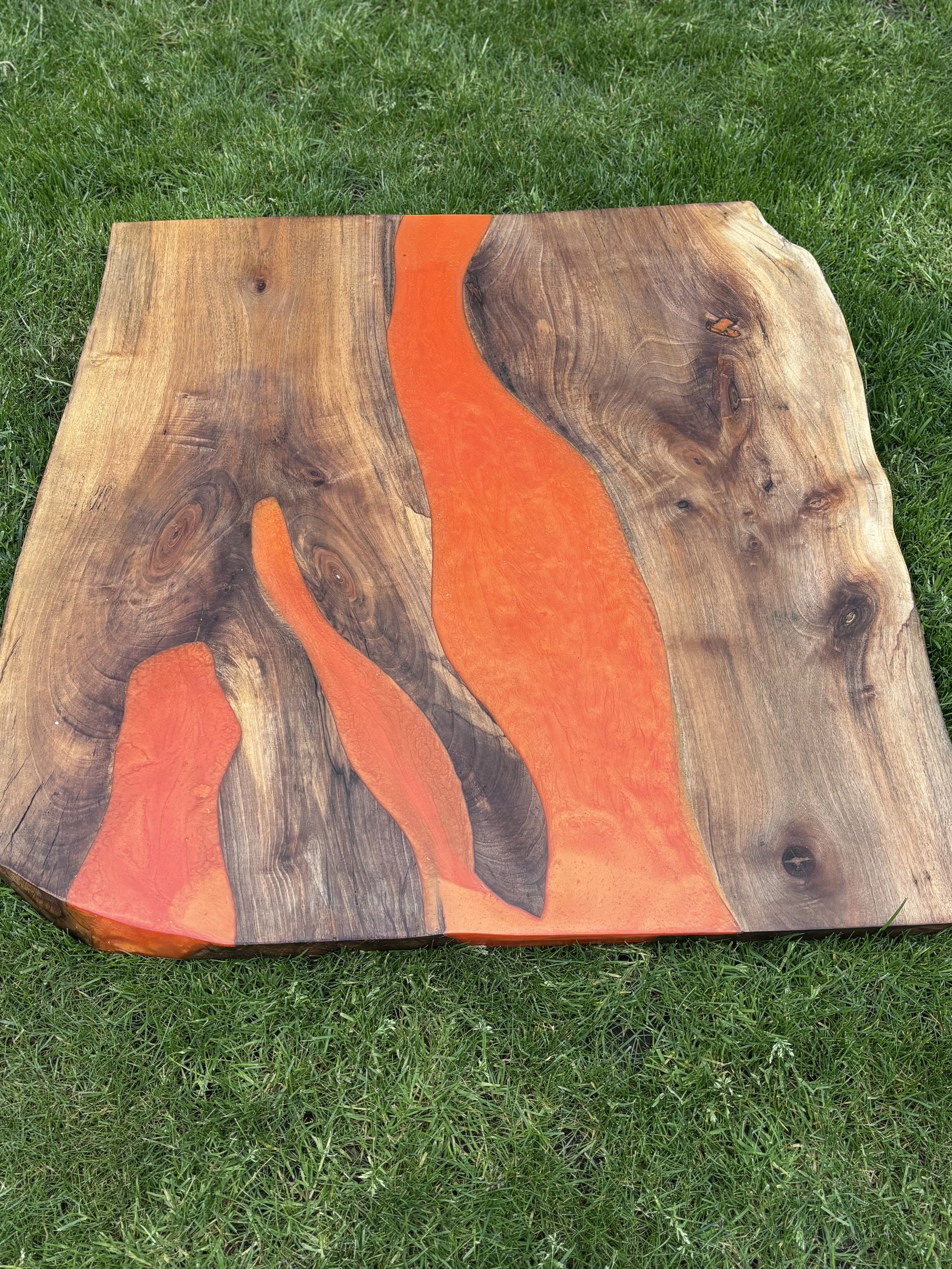 A wooden table with a natural edge, featuring inlaid orange resin in a flowing, wavy pattern that mimics a river.