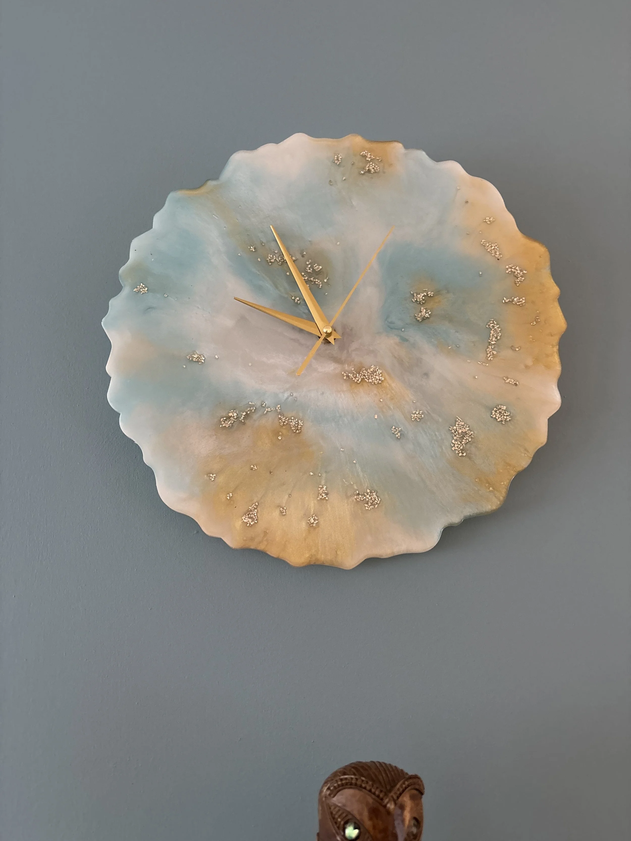 A decorative wall clock with a shell or coral shape featuring light pastel colors and gold accents, mounted on a blue wall.
