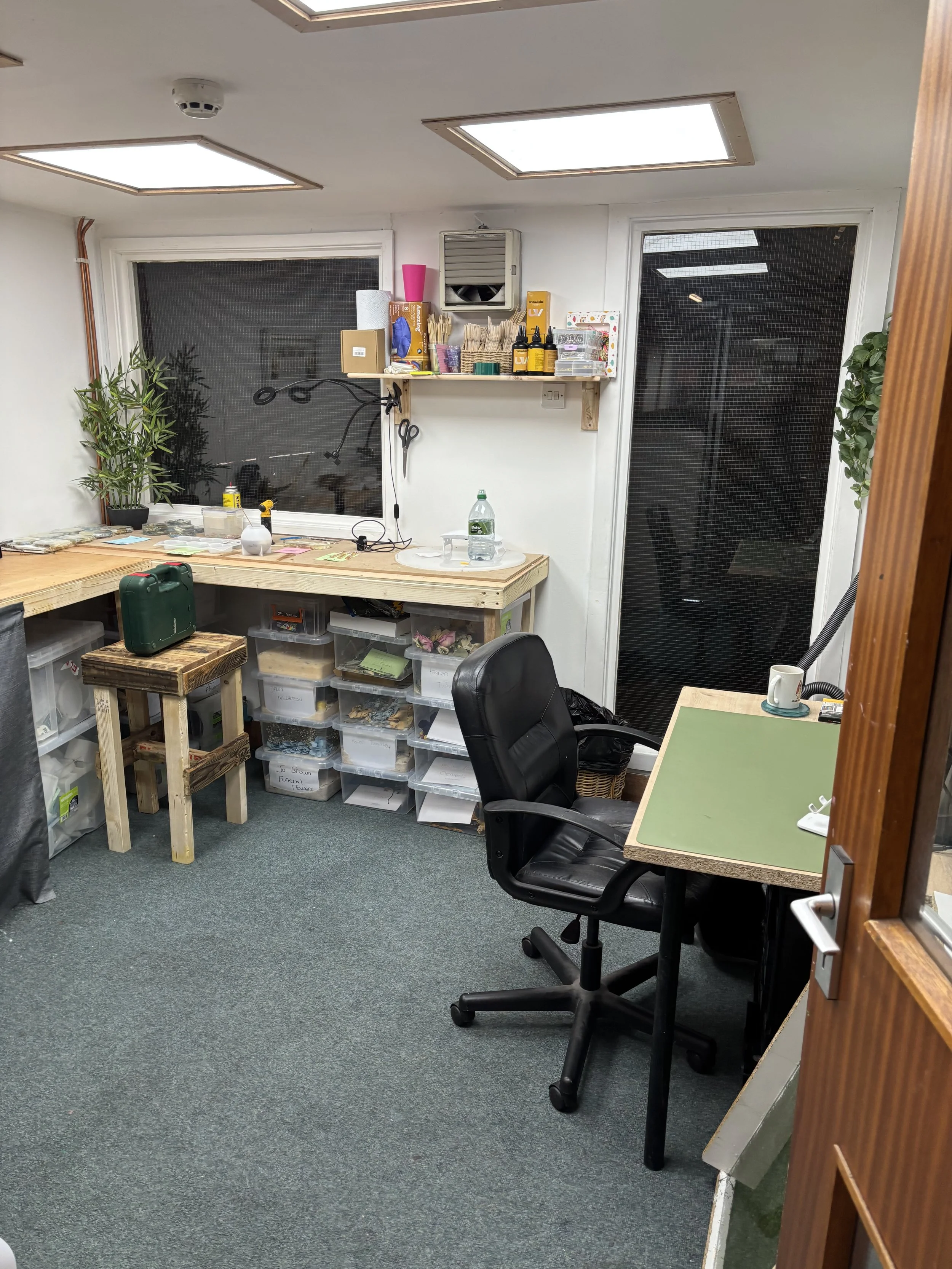 A cluttered office or workshop with a wooden table, plastic storage bins underneath, a black office chair, and a green work surface. The back wall has two windows and a small shelf with various tools and supplies, including bottles, scissors, and paper. There is a plant on the left side of the window and a coffee mug on the right side of the work surface.