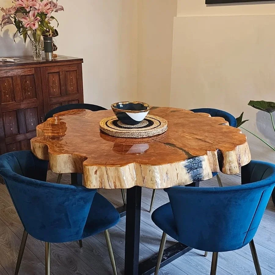 A wooden dining table with a natural, live edge, surrounded by four blue chairs with gold accents. On the table, there is a black and white ceramic bowl on a woven placemat. In the background, a wooden sideboard with a bouquet of pink lilies and some