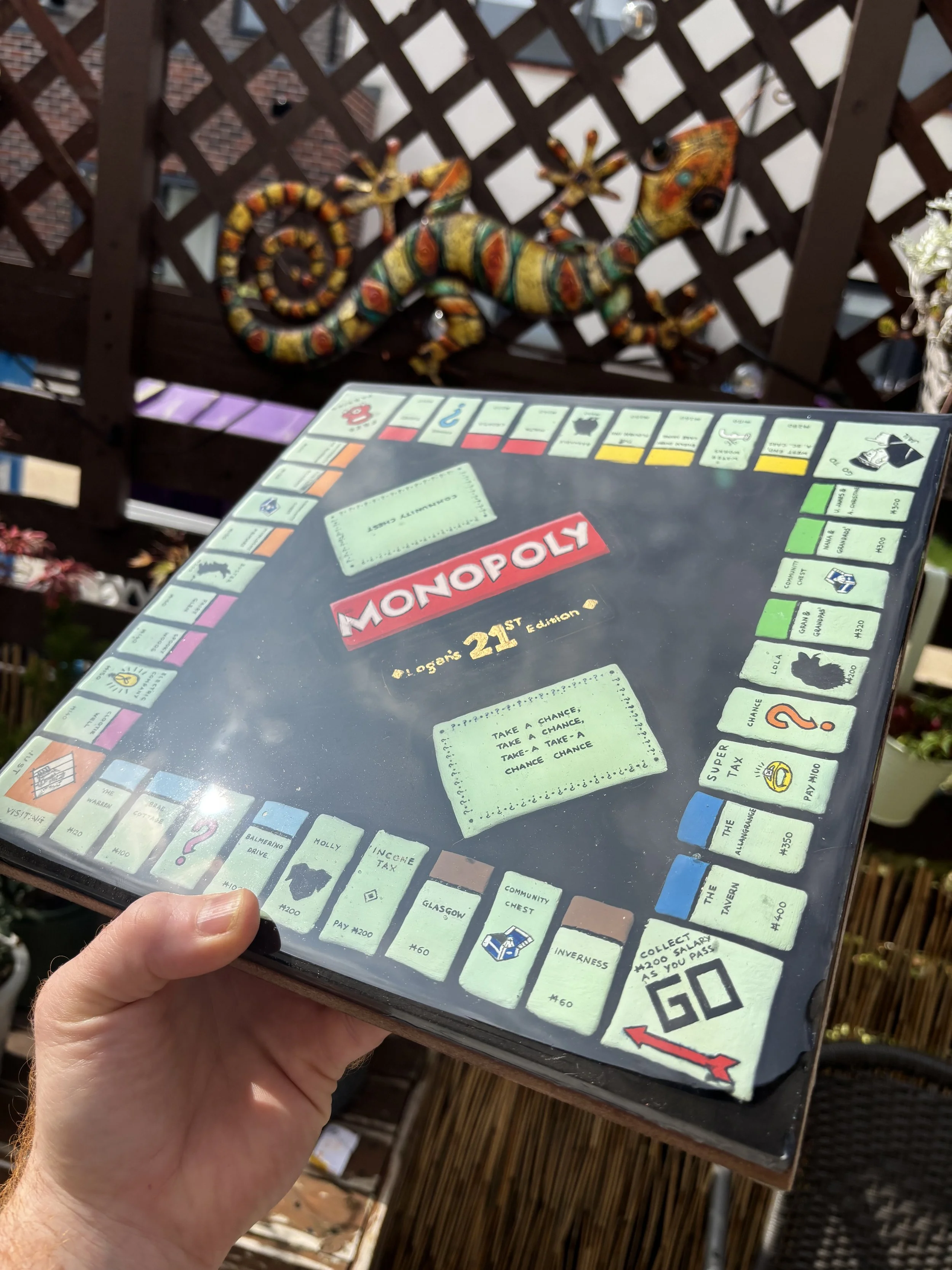 Close-up of a person holding a Monopoly board game outside on a patio with wooden lattice and a colorful ceramic lizard decoration in the background.