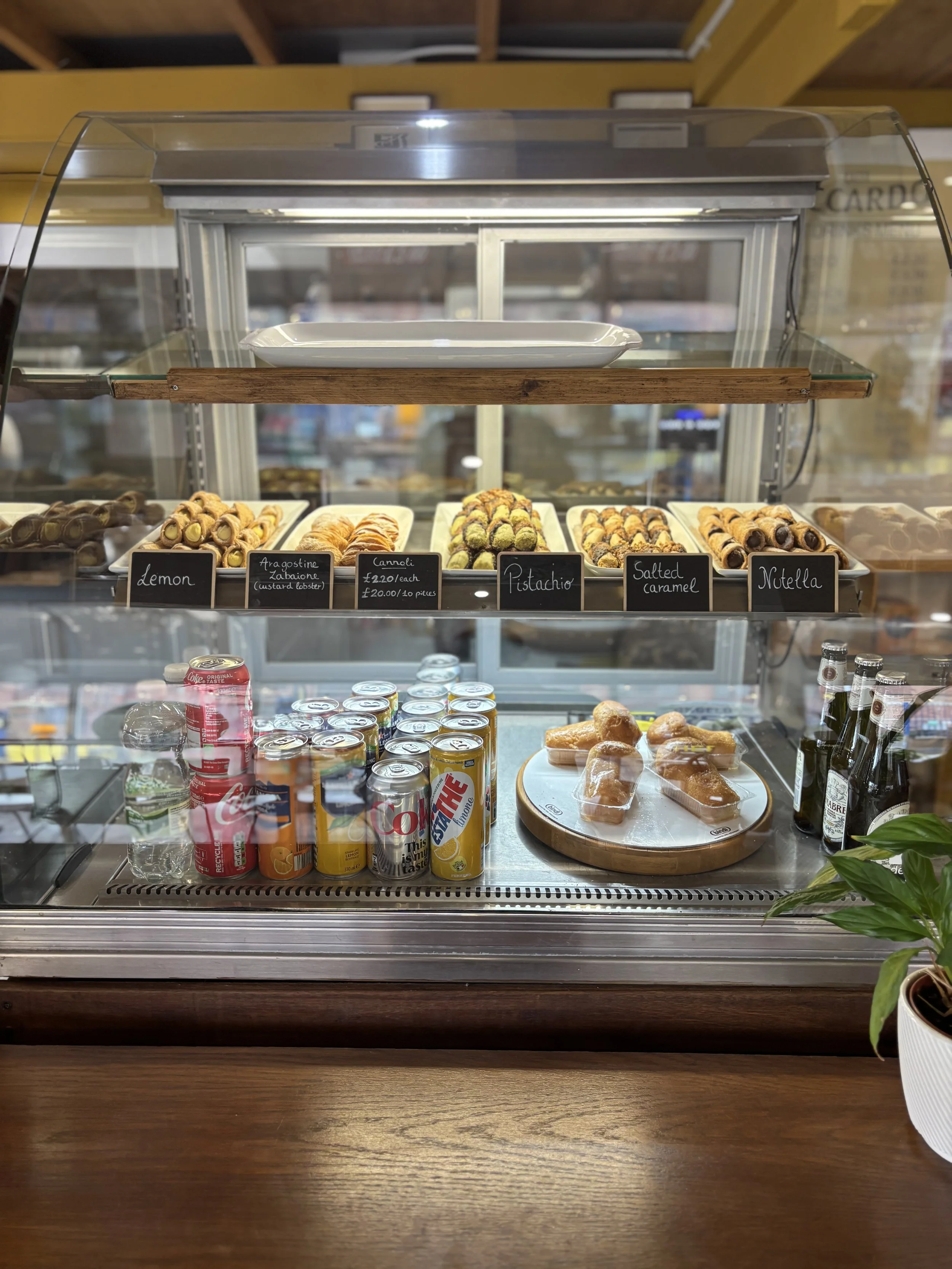Display case with various types of sushi rolls, labeled with small black signs, and cans of soda on a wooden counter.