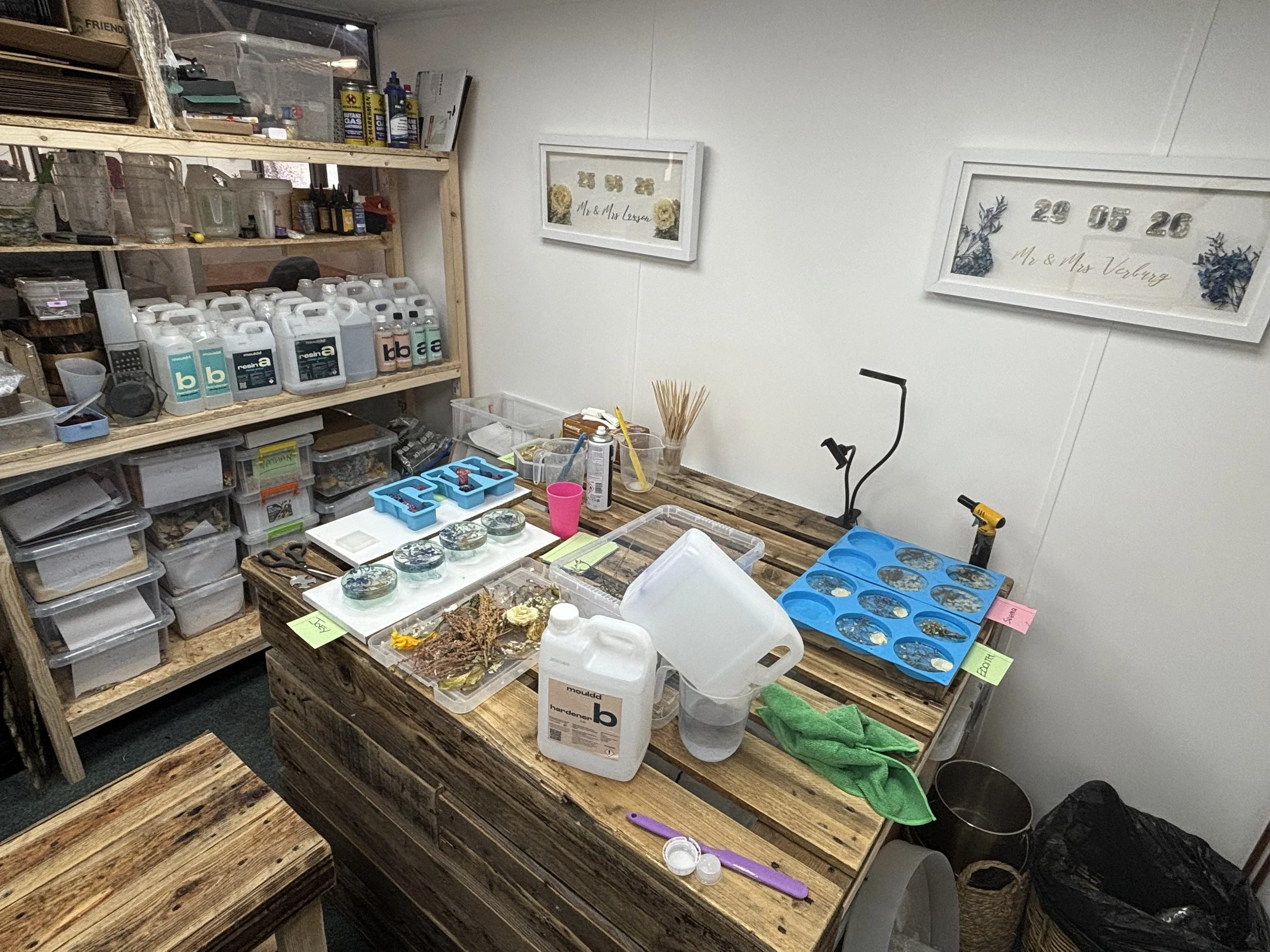 Workshop with wooden tables, shelves with supplies, framed artwork on the wall, and various tools and containers on the tables.