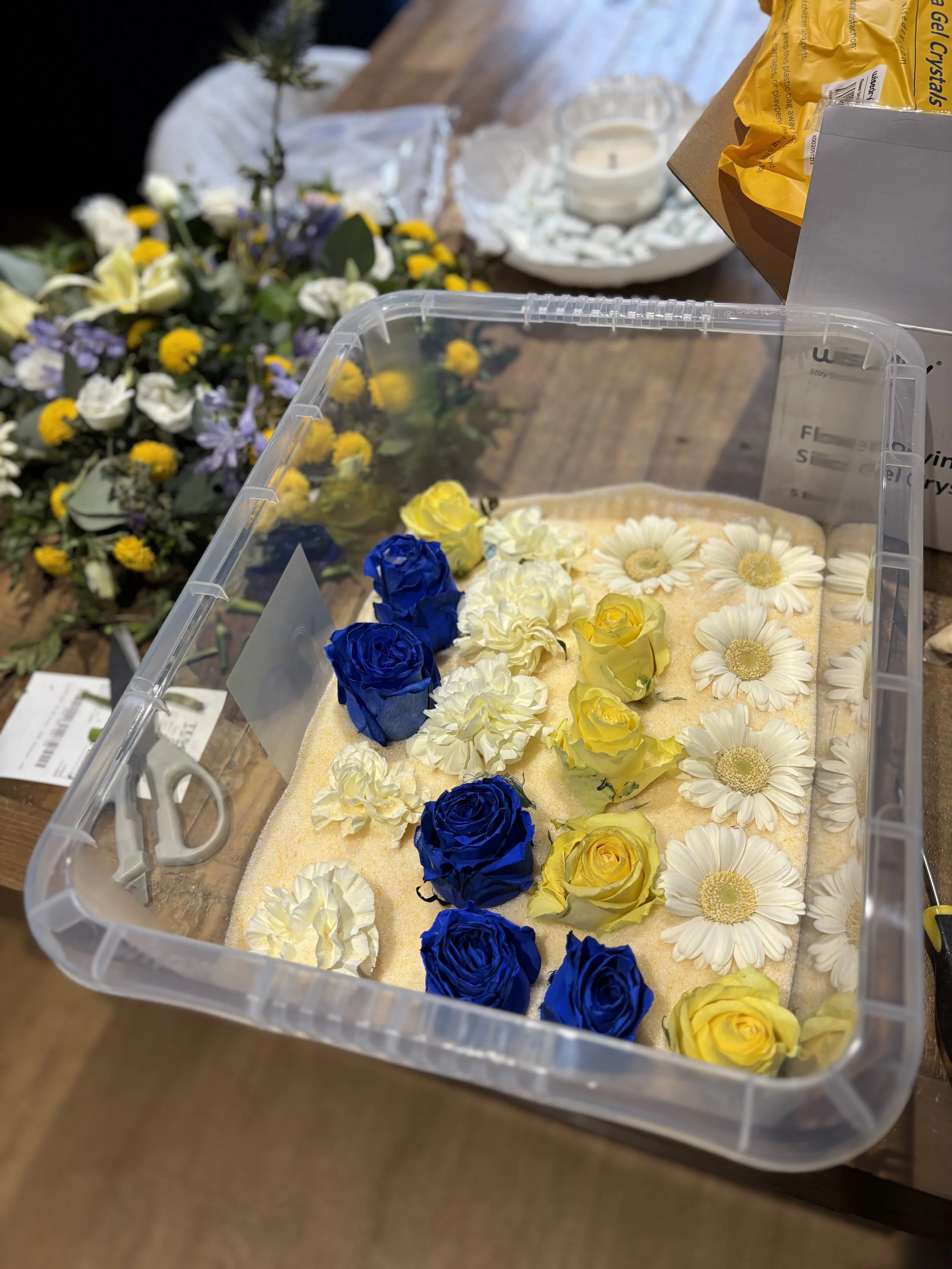 A clear plastic container filled with various blue, yellow, and white flowers, placed on a wooden table.