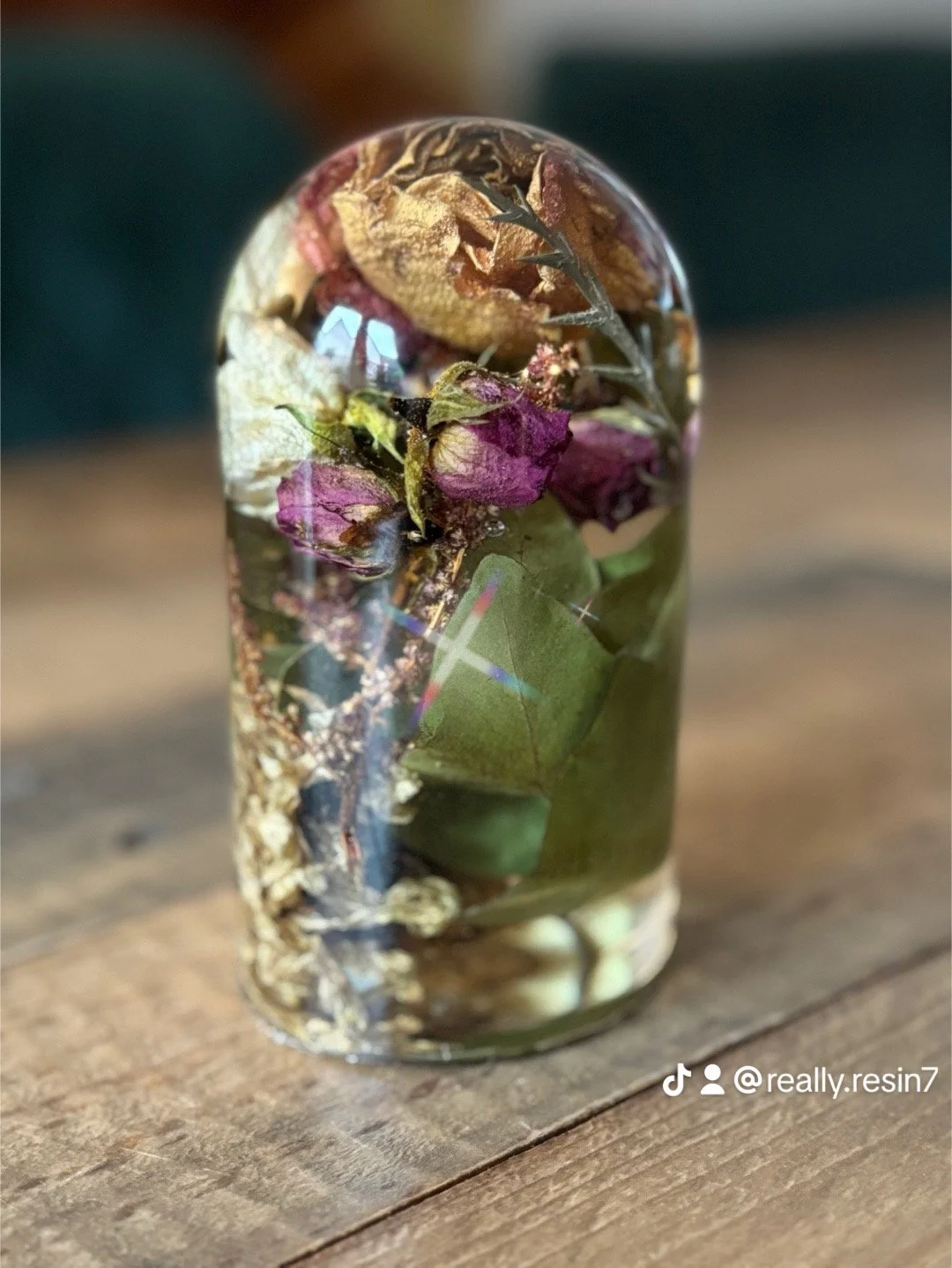 A small glass jar filled with dried flowers and leaves, including purple and green foliage, placed on a wooden surface.